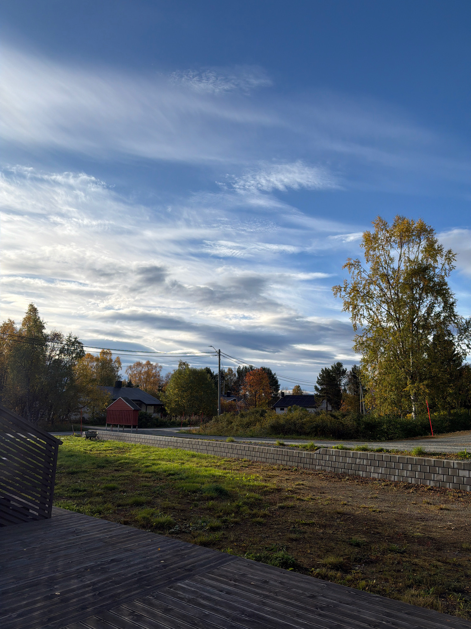 A full, outdoor, vertical shot depicts a residential area in Storsteinnes, Norway, under a bright, partly cloudy sky. The foreground features a dark wooden deck with a slatted railing. Beyond the deck, a grassy area leads to a paved pathway bordered by a low, stacked stone wall. A red, shed-like structure with a dark roof sits to the left of the pathway, adjacent to a small red building. In the background, several houses are visible among trees displaying autumn foliage in shades of yellow and orange. A tall tree with yellowing leaves stands prominently on the right side of the frame. A street with parked cars is visible in the mid-ground, and power lines stretch across the scene. The overall impression is one of a quiet, suburban autumn day.