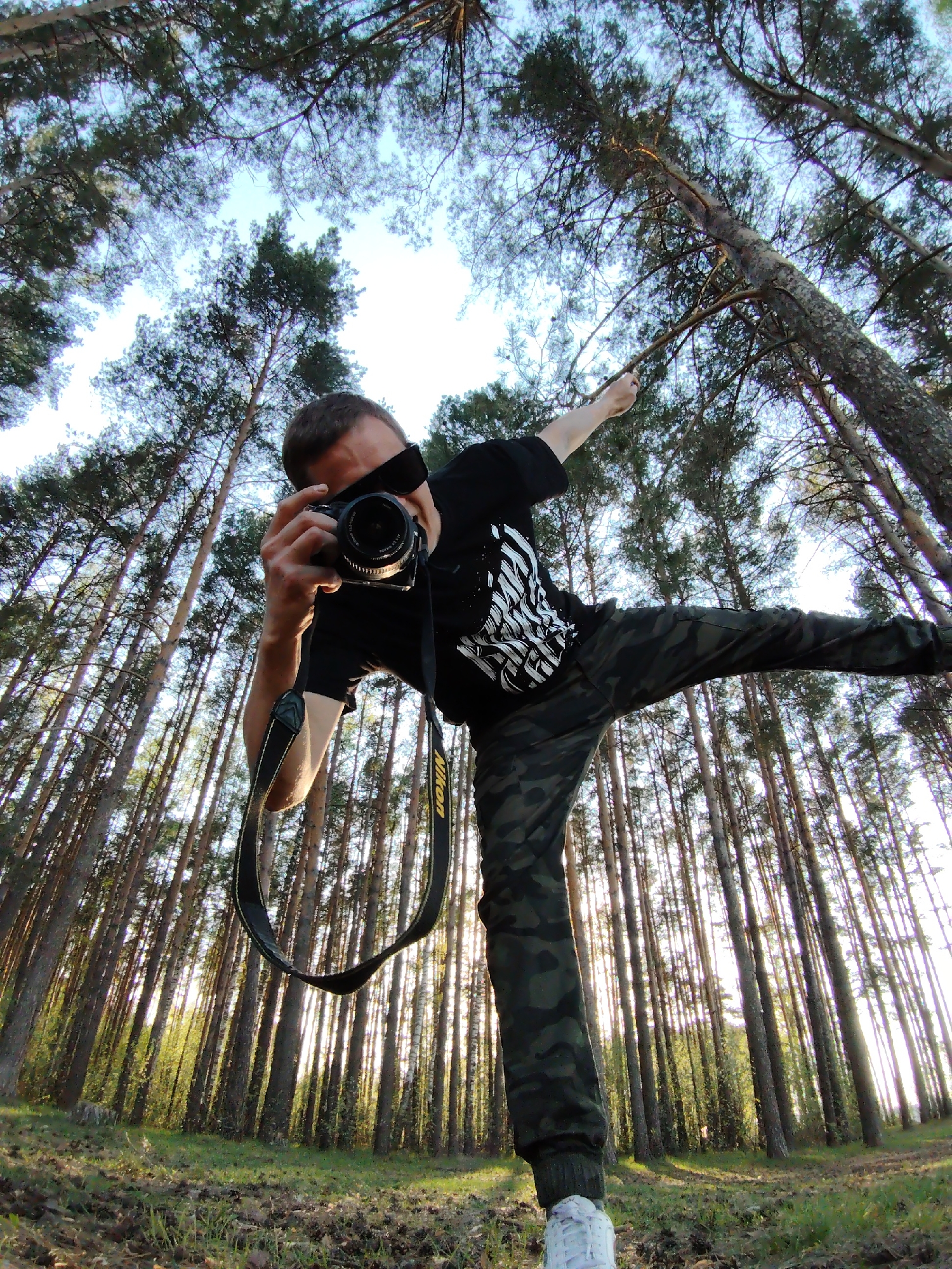 A low-angle shot captures a man in a forest setting. The man is wearing sunglasses, a black t-shirt with a graphic, camouflage pants, and white shoes. He is holding a Nikon camera with a lens and a black Nikon strap in front of him, pointing it towards the viewer as if taking a picture. His right leg is extended outwards, and his right arm is raised, gesturing towards the trees. The forest is characterized by tall, slender trees that reach towards the sky, creating a perspective that emphasizes height. The ground is covered in grass and fallen leaves. The sky is visible through the treetops, appearing light and bright. The location is Chaikovsky, Russia.