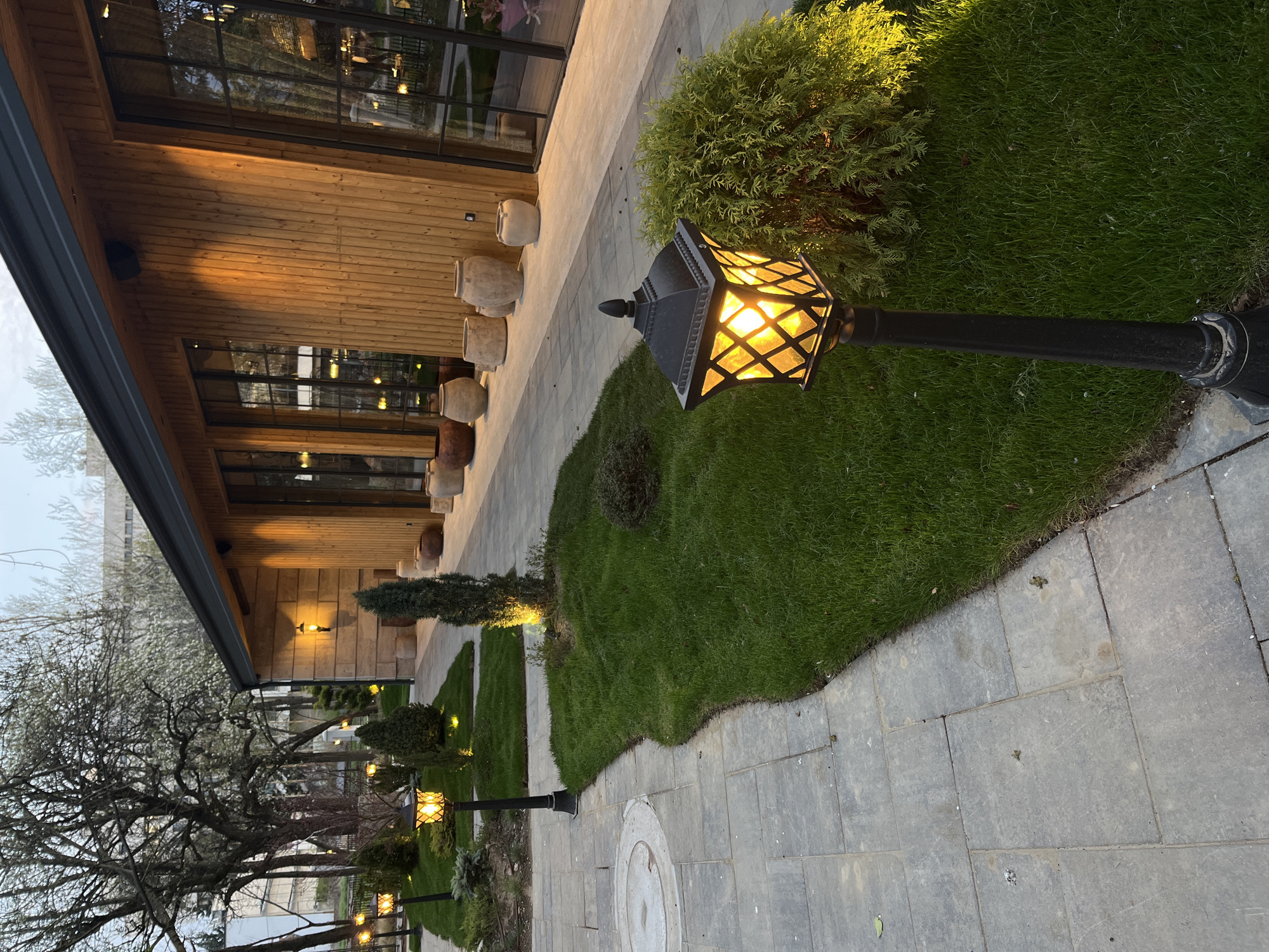 The image presents a tranquil outdoor scene at dusk or early evening, likely in Sofia, Bulgaria. A paved pathway of gray rectangular stones winds through a meticulously landscaped area, running parallel to a modern building. The building features a natural wooden facade with horizontal planks and large, dark-framed windows that reflect the subtle light of the sky and nearby trees. A series of large, light-colored ceramic pots decorates the area directly outside the building.
The pathway is lined with lush green grass areas, interspersed with neatly trimmed bushes and smaller trees. Several black, traditional lantern-style street lamps are a prominent feature, casting a warm yellow glow across the scene. One lamp stands in the immediate foreground, its bright light illuminating a dense green bush and a section of grass. Further along the path, additional similar lamps can be seen, providing ambient lighting. In the background, larger, bare-branched trees, some hinting at early spring blossoms, complete the natural setting.
No people or activities are visible, contributing to the peaceful atmosphere. The artificial lights suggest a time of day when natural light is fading, enhancing the cozy and inviting feel of the area. No visible text is present in the image.
