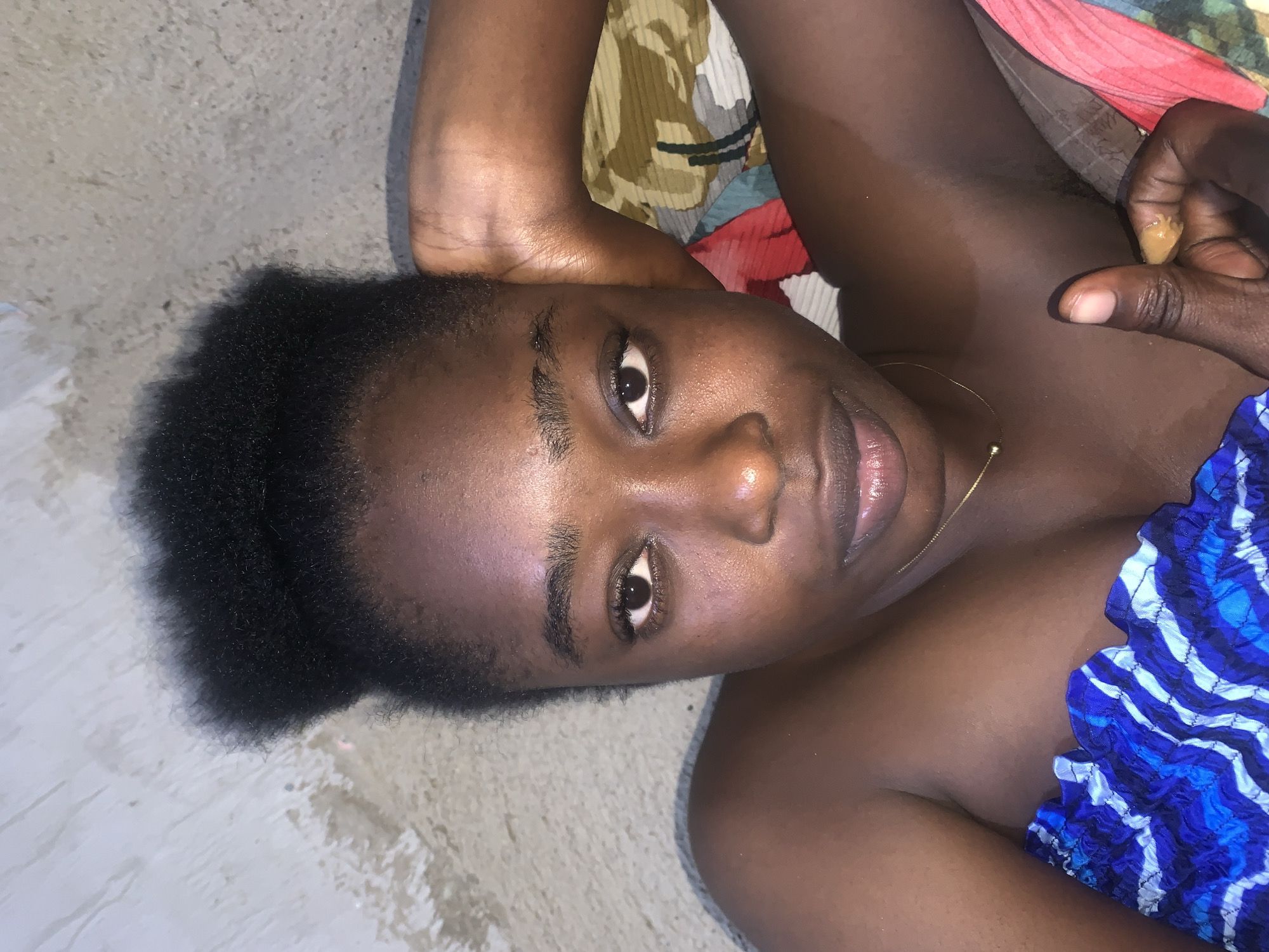 A young Black woman lies on her back, looking upwards towards the camera. Her dark, curly hair is pulled up into a high bun. She wears a blue and white patterned top, and a thin gold necklace. Her right arm is bent, with her hand resting on the back of her head, and her left hand is visible giving a thumbs-up gesture. She is on a bed with a patterned sheet. The background appears to be a wall and a floor, suggesting an indoor setting. The image is likely taken in the city of Rafin Par, Nigeria, as indicated by the prompt.