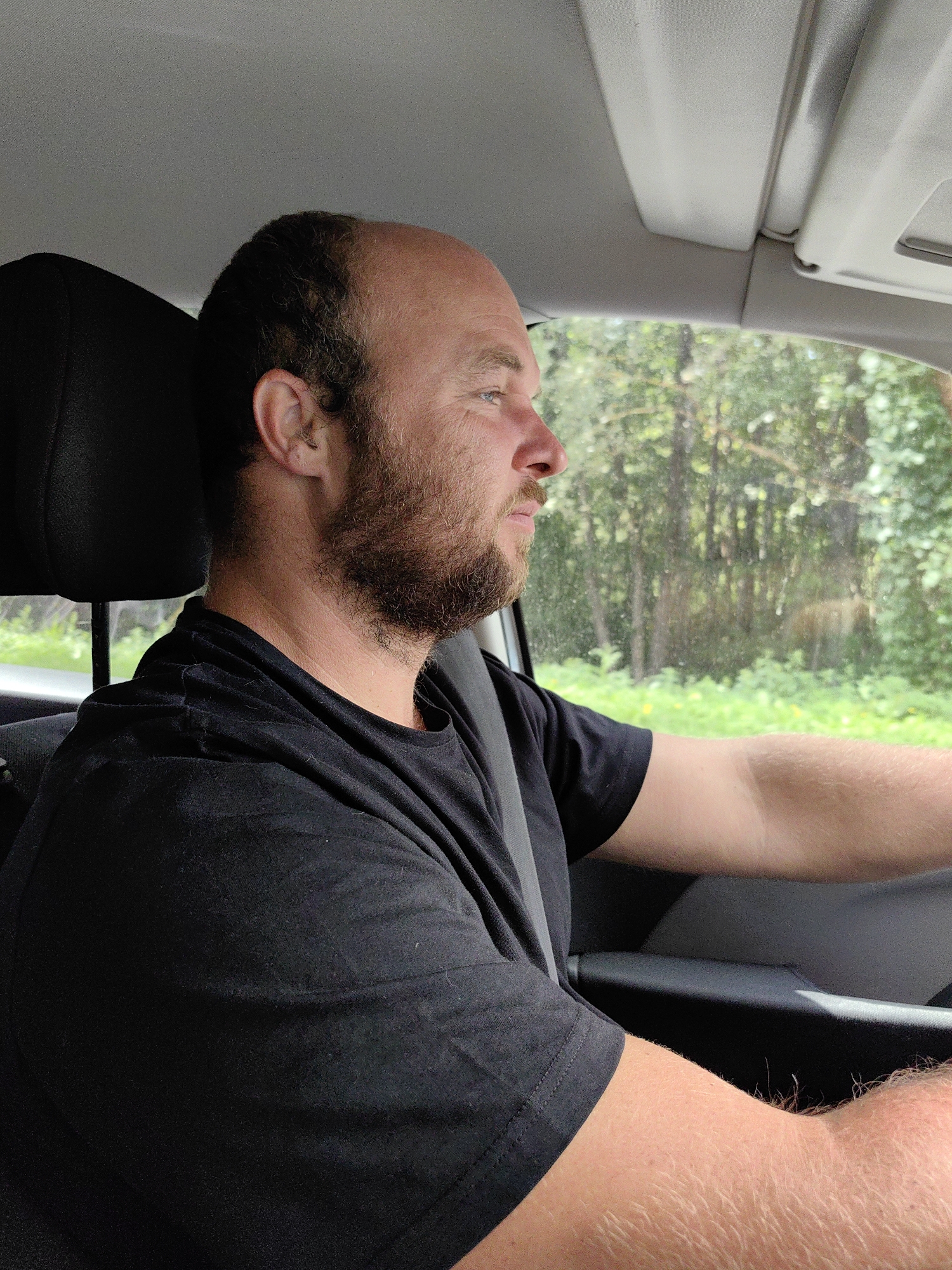 An adult male with a receding hairline and beard is driving a car. He is wearing a black t-shirt and a seatbelt. The car's interior is visible, along with trees and greenery outside the window. The scene is consistent with a rural road in the Мядзельски сельскі Савет, Belarus, likely during daylight hours.