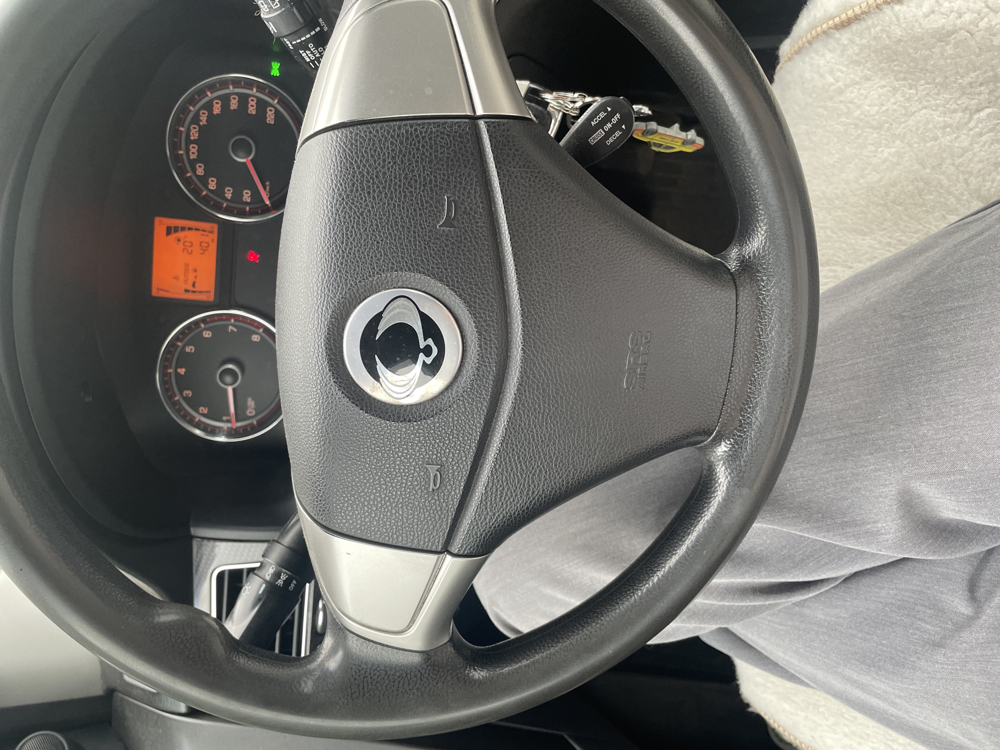 The image shows a close-up view of the interior of a car, specifically the steering wheel and dashboard. The steering wheel has a black leather grip and silver accents. In the center of the steering wheel is a circular emblem with a stylized logo. Below the emblem, "SRS AIRBAG" is visible. To the left of the steering wheel, a turn signal stalk with controls for lights is partially visible. On the dashboard, a digital display shows "OUTSIDE 20" and "40" with some graphics, next to a speedometer that reads up to 220 km/h. To the right of the speedometer is another stalk, likely for windshield wipers, with settings like "MIST," "OFF," "AUTO," and "SLOW." A set of car keys with a fob labeled "CRUISE ON-OFF," "ACCEL ▲," and "DECEL ▼" is attached to the steering column. The lower part of the image shows a person's legs covered in gray pants, and a white fluffy seat cover. The scene is lit by the car's interior lighting, with an orange glow from the dashboard display. No information about the city of Gavrilovka, Russia, can be derived from the image.