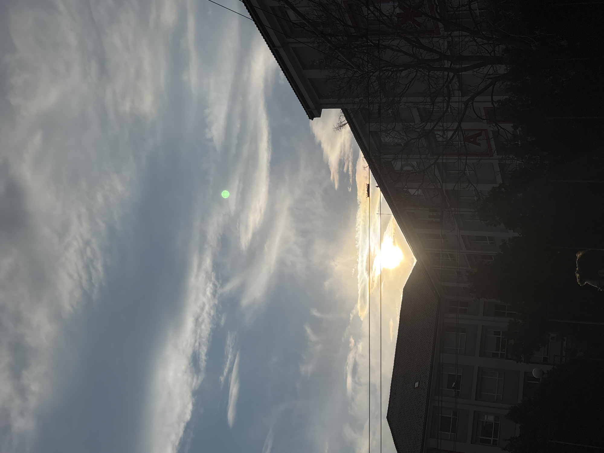 This image, taken in Varna, Bulgaria, depicts an urban sky scene likely during late afternoon or early morning, characterized by strong backlighting. The sky fills most of the frame, dominated by a mix of light blue-grey tones and elongated, wispy white clouds.
A bright, golden sun is positioned in the mid-right portion of the image, partially obscured by a building, creating a distinct triangular flare of light. A prominent green lens flare is visible in the upper-left area of the sky.
Two buildings are sharply silhouetted against the bright sky and sun. One building on the right has a sloped, tiled roof. Another, taller building, is visible in the lower-center-right, showing multiple stories and faint outlines of windows. A small, white satellite dish is identifiable on this taller structure. Dark, bare tree branches extend into the frame, intertwining in front of and between the buildings, further obscuring their details. Thin power lines or cables are also visible stretching across the scene.
No people are present in the image. The strong contrast between the illuminated sky and the dark silhouettes of the urban elements creates a dramatic and contemplative mood. On the darker building, a faint, large red letter, possibly part of a sign, is discernable through the tree branches.