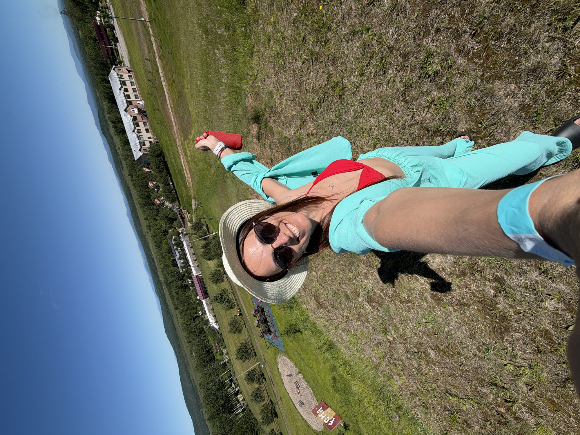 A woman stands on a grassy hill, taking a selfie. She wears a red bikini top, a turquoise cover-up, a wide-brimmed hat, and sunglasses. In her right hand, she holds a red portable speaker. She is on a slope overlooking a valley with trees, buildings, and mountains in the distance. The sky is clear and blue. The area appears to be a park or recreational area, possibly part of a resort. The sign "FOMA" is visible on the ground to her left. The landscape suggests a rural or semi-rural location outside of a major urban center. Based on the context of previous images, this location is identified as being near Birobidzhan, Russia.