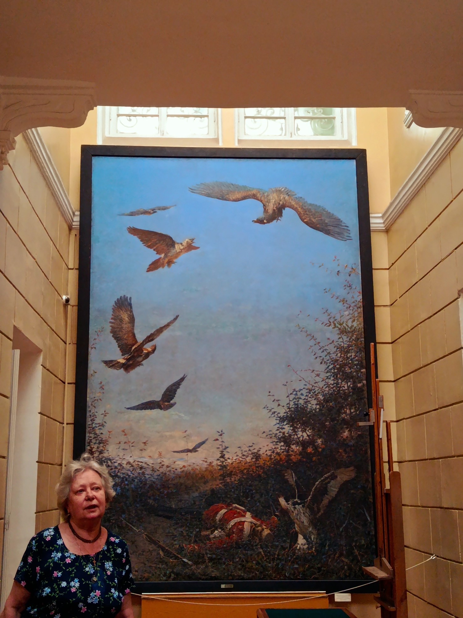 A medium shot captures a woman standing to the left of a large, framed painting. The woman, with light blonde hair and fair skin, wears a dark blue floral print dress and a dark necklace. Her gaze is directed slightly upwards and to the right.
The painting, which dominates the frame, depicts a flock of birds in flight against a clear blue sky. Several birds are visible, some in mid-flight with outstretched wings. The bottom portion of the painting shows a wooded area with foliage and a hint of red fabric on the ground. The painting is displayed on an easel.
The background features an interior wall with a textured finish, possibly stone or stucco, in a warm yellow-beige color. Decorative architectural elements, such as ornate moldings and a pediment, are visible at the top of the frame. There is a security camera mounted on the wall to the left of the painting. Two small windows with decorative grilles are situated above the painting. The lighting suggests an indoor environment.