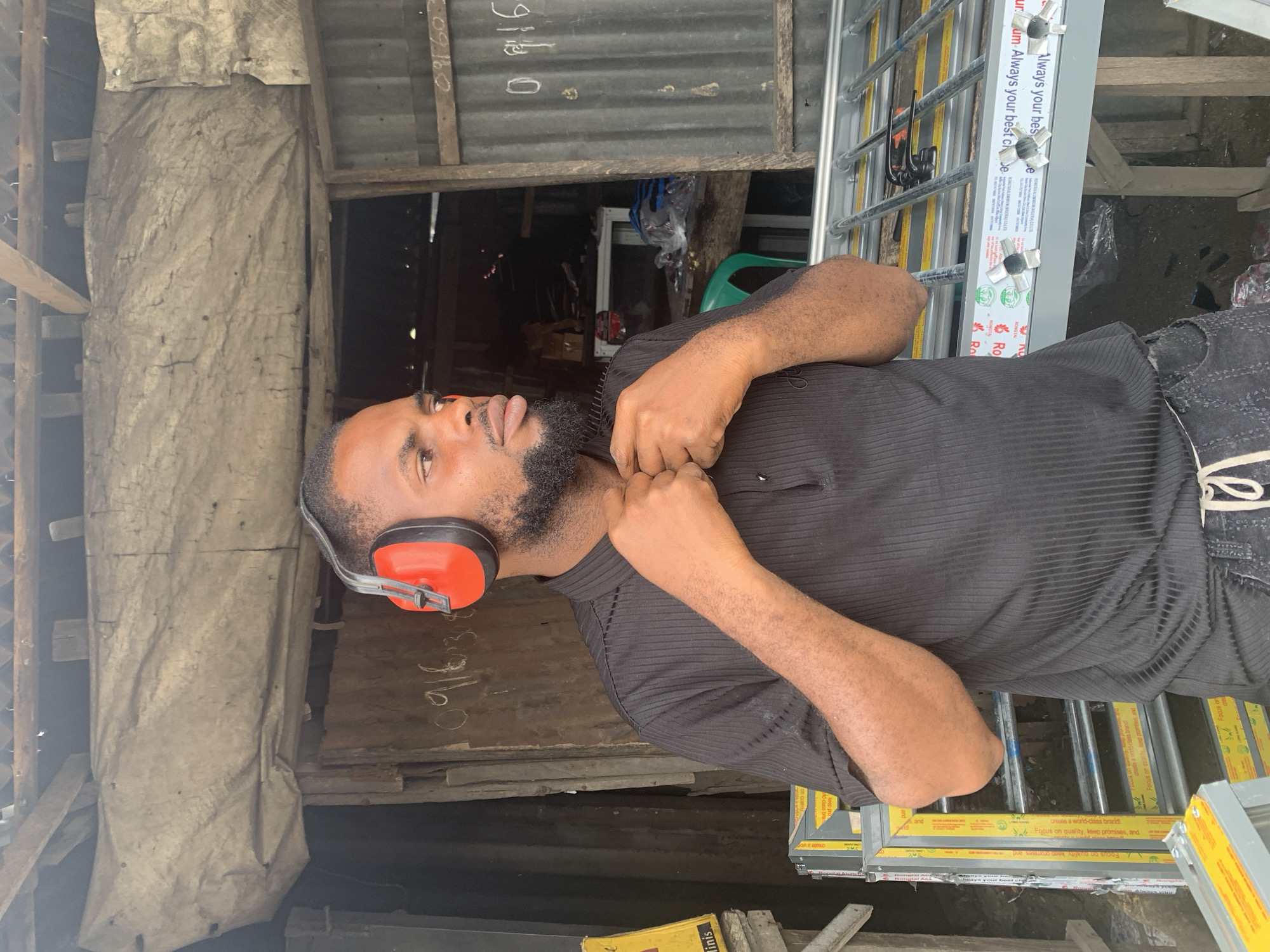 A man is standing in what appears to be a workshop or construction site, possibly in Umuode, Nigeria. He is wearing a black, ribbed polo shirt and dark jeans, with orange earmuffs over his ears. His right hand is resting on his neck, and his left hand is partially visible. In the background, there are corrugated metal sheets, wooden planks, and what looks like a tarp hanging on the left. To the right, there are several aluminum window frames stacked up. The window frames have yellow labels with text, including "Always your best cie" and "Focus on quality, keep promises, and create a world-class brand!". There is also some handwritten text on the corrugated metal, including "0916". The scene is lit by natural light, suggesting it is daytime. The man's expression is neutral.