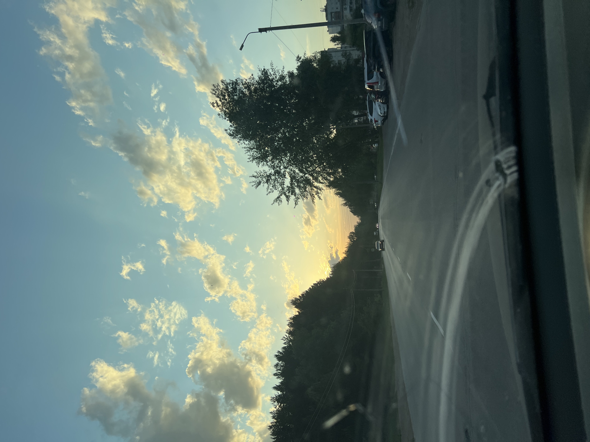 The image depicts a view from inside a vehicle traveling along a road, possibly in the city of Chaikovsky, Russia. The sky is visible with light clouds, indicating a time around sunset or sunrise, as the light is soft. Trees line the road, and there are parked cars on the right side of the road. A streetlight is also present.