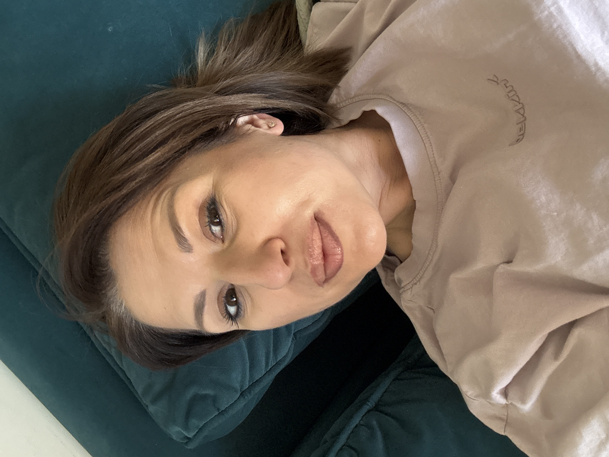 A woman with brown hair and light skin lies on a green velvet couch. She is wearing a light brown sweatshirt with a small, curved inscription on the upper right chest area. Her head is tilted slightly, and she is looking directly at the camera. No specific location in Sofia, Bulgaria, is identifiable from the image.