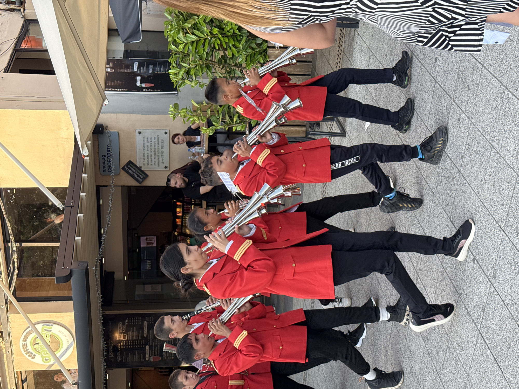 The image depicts an urban outdoor scene on Svobodata Square in Pleven, Bulgaria, under bright daylight. A group of approximately eight to ten young individuals, appearing to be part of a marching band, are walking from left to right across the foreground. They are uniformly dressed in red single-breasted jackets with gold-colored trim on the cuffs, black trousers, and various athletic sneakers. Each individual is holding and playing a musical instrument; most are silver-toned brass instruments resembling trumpets or bugle-like fanfare instruments, while one individual towards the left is playing a clarinet.
In the background, on the left, is a storefront with a dark overhang. A circular green logo for "Coffee Point Garden" is visible through a large window, behind which a female individual can be seen. Adjacent to this, a tall dark menu board with Cyrillic text and beverage images is positioned outside another establishment.
Towards the center-right background, an outdoor cafe seating area is visible beneath a large, cream-colored retractable awning. Two adult individuals, a bearded male in a black t-shirt and a female in a dark top, are seated at a wooden table with drinks, partially obscured by green potted plants. Above this area, affixed to a building column, is a blue sign with white Cyrillic text "ПЛОЩАД СВОБОДАТА" (Svobodata Square). Below this, a dark commemorative plaque with white Cyrillic text is also visible.
In the immediate foreground on the right, the back and right profile of a female individual wearing a black and white vertically striped sleeveless dress is visible, with light brown hair. The ground surface is paved with light gray square or rectangular concrete tiles.