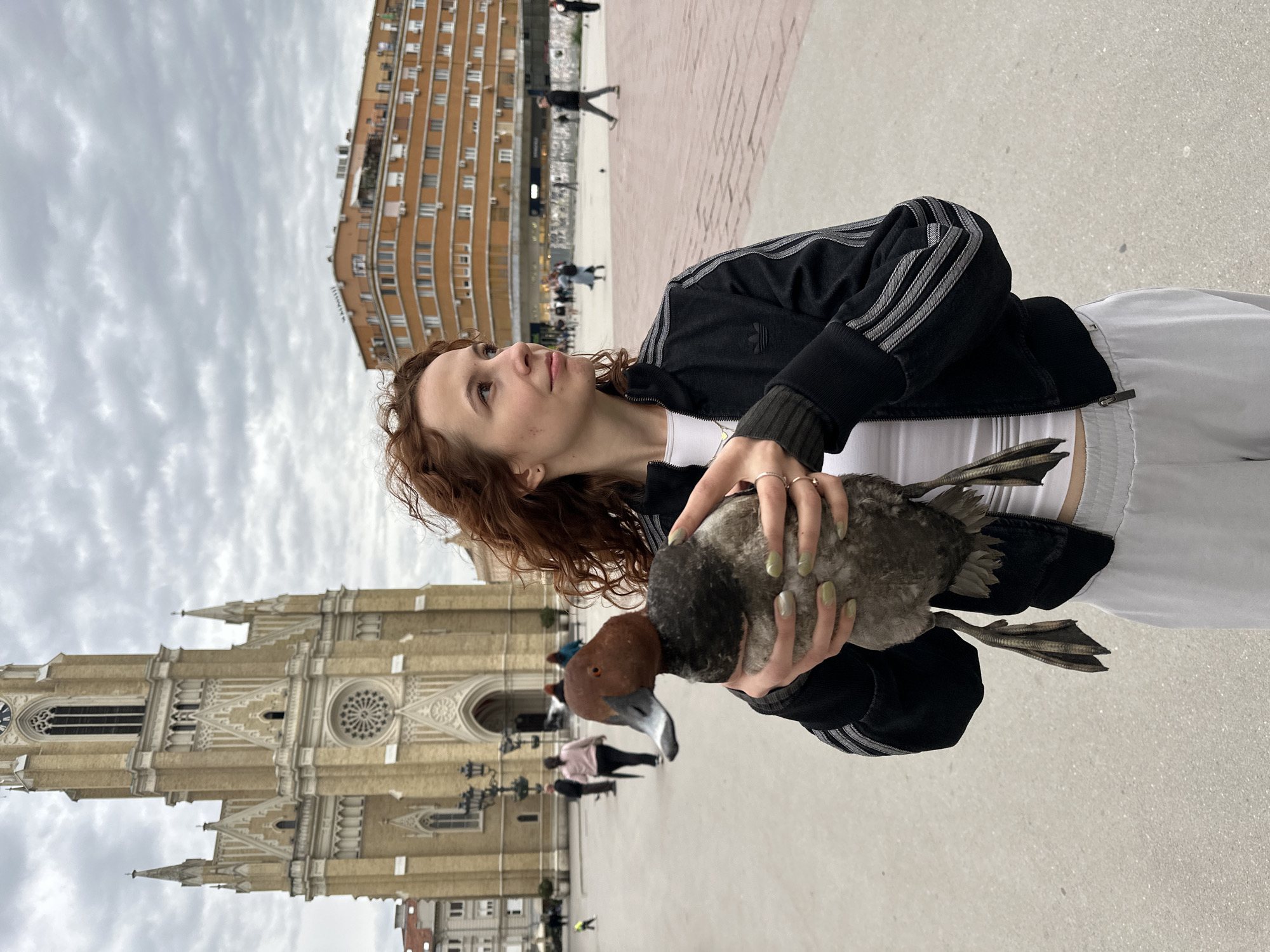 A young woman is captured in a public square in Novi Sad, Serbia, holding a duck under an overcast sky. She has reddish-brown curly hair and is wearing a black track jacket with white stripes on the sleeves, a white top, and light-colored pants. Her fingers bear several rings, and her nails are painted light green. She cradles a duck with both hands; the duck has a reddish-brown head, a pale beak, and a grayish body with visible webbed feet. The woman looks upwards and slightly to her left with a calm expression.
In the background, to the left, stands a large, ornate Gothic-style building, identifiable as the Name of Mary Church (also known as the Cathedral), featuring tall spires, pointed arches, and a prominent rose window. To the right in the background, a multi-story, curved building with numerous windows and balconies rises, characteristic of urban architecture. The ground is a light-colored paved surface, indicating a spacious pedestrian area. Several indistinct figures of people are visible walking in the distance, suggesting a bustling public space. The sky is largely covered with clouds, indicating an overcast day. There is no visible text in the image.