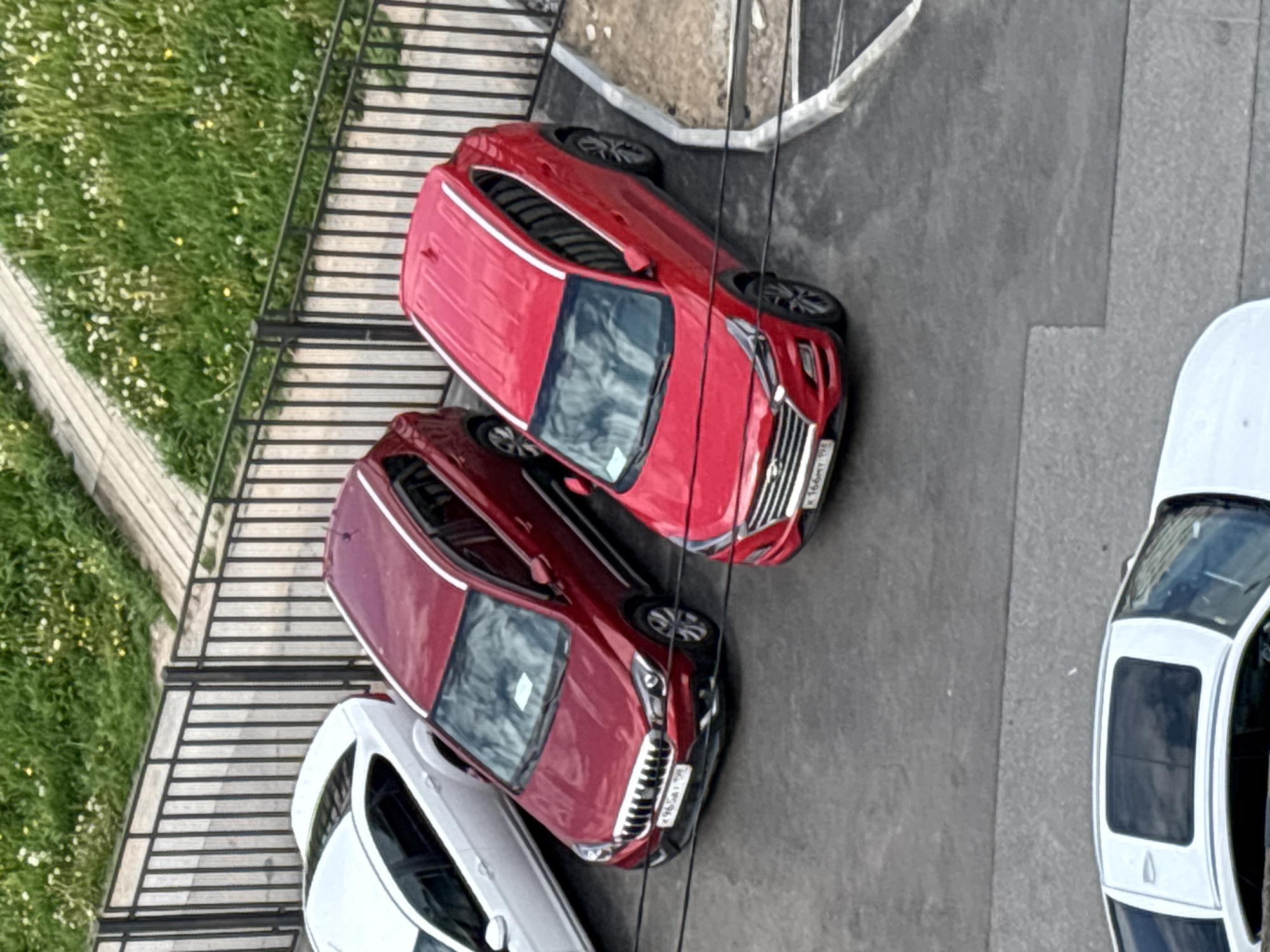 The image, taken from a high angle, captures a parking area adjacent to a grassy patch, likely in Saint Petersburg, Russia. The scene features four parked cars: three red/maroon compact SUVs/crossovers and one white car, also appearing to be a compact SUV or hatchback, partially visible on the far right.
The top-most visible red car has the license plate "K 166 MY 198". The red car in the middle has the license plate "K 965 AM 198". The "198" region code on these plates confirms the location as Saint Petersburg and the Leningrad Oblast.
To the left of the cars is a dark metal fence or railing, bordering a vibrant green lawn dotted with small white and yellow wildflowers. Beyond the fence, a lighter-colored, possibly wooden, walkway or terrace is visible. The cars are parked on a grey paved surface, likely asphalt or concrete.
The image depicts an ordinary daytime scene, with natural, diffused lighting suggesting either an overcast sky or mid-day sun. No people or specific activities are visible; the focus is on the parked vehicles and their immediate surroundings.