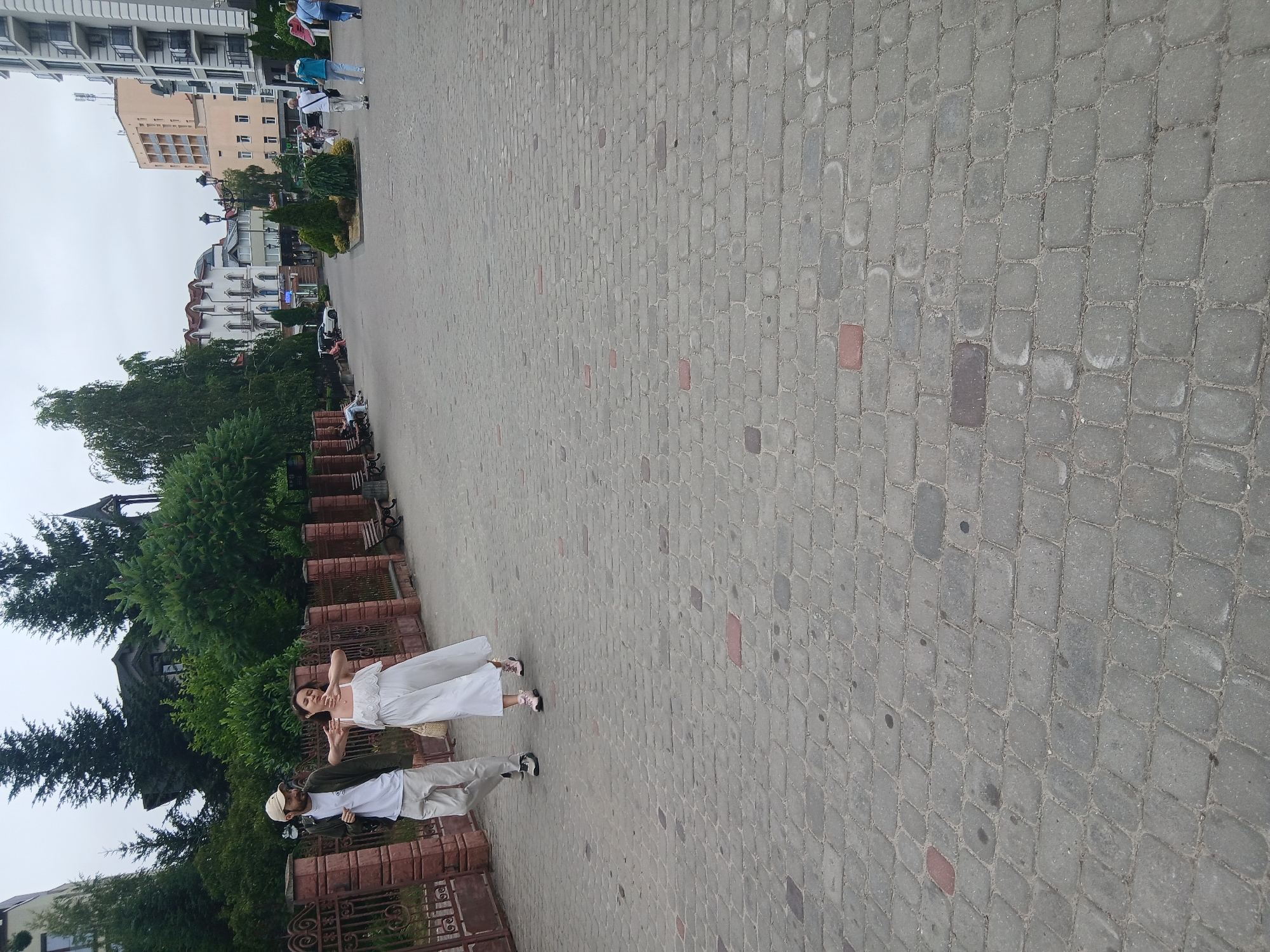 The image shows a street scene in Truskavets, Ukraine. Two people are walking on a cobblestone path. The person in front is a woman wearing a white dress and light pink shoes. The person behind her is a man wearing a white t-shirt, light-colored pants, a dark jacket, a baseball cap, and dark shoes. They appear to be walking away from the camera, towards a row of buildings and lush green trees.
The background includes a multi-story building with balconies and arched windows, as well as other buildings with red-tiled roofs. The sky is overcast, suggesting a cloudy day. The cobblestone pavement is made of grey rectangular stones, with some red and dark grey stones interspersed. A decorative metal fence with intricate scrollwork lines the left side of the path.
There are no visible signs with text. The overall atmosphere appears to be leisurely, possibly a stroll through a town or resort area. The subjects' postures suggest they are engaged in conversation or simply enjoying a walk. The time of day is likely daytime, given the brightness, but the overcast sky prevents strong shadows.