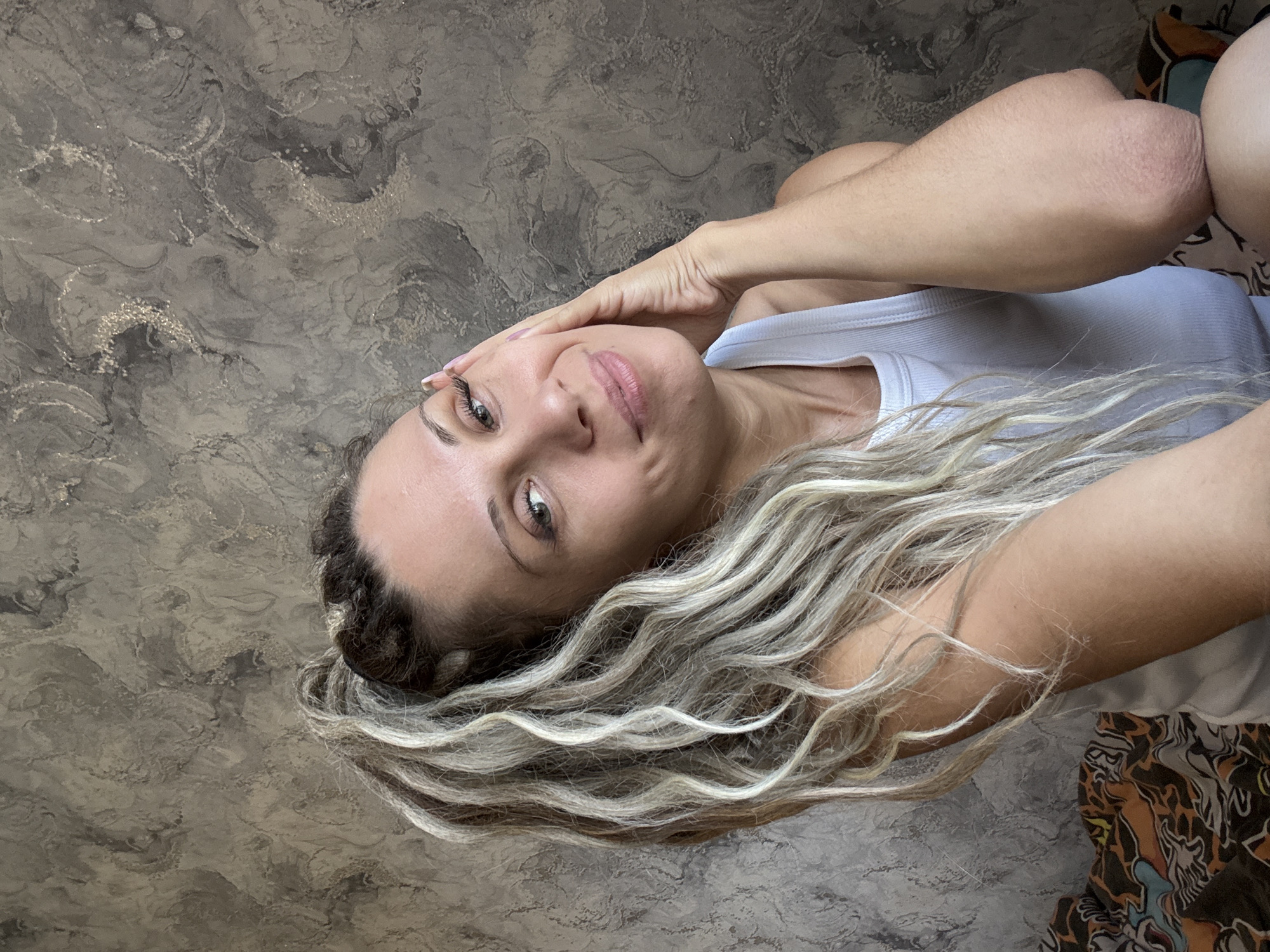 In Rusanovo, Russia, a woman with blonde, curly dreadlocks tied in a high ponytail poses for a selfie against a textured gray wall. She wears a white tank top and rests her right hand on her cheek, looking towards the camera with a slight smile. The background features a patterned blanket on the left side of the frame.