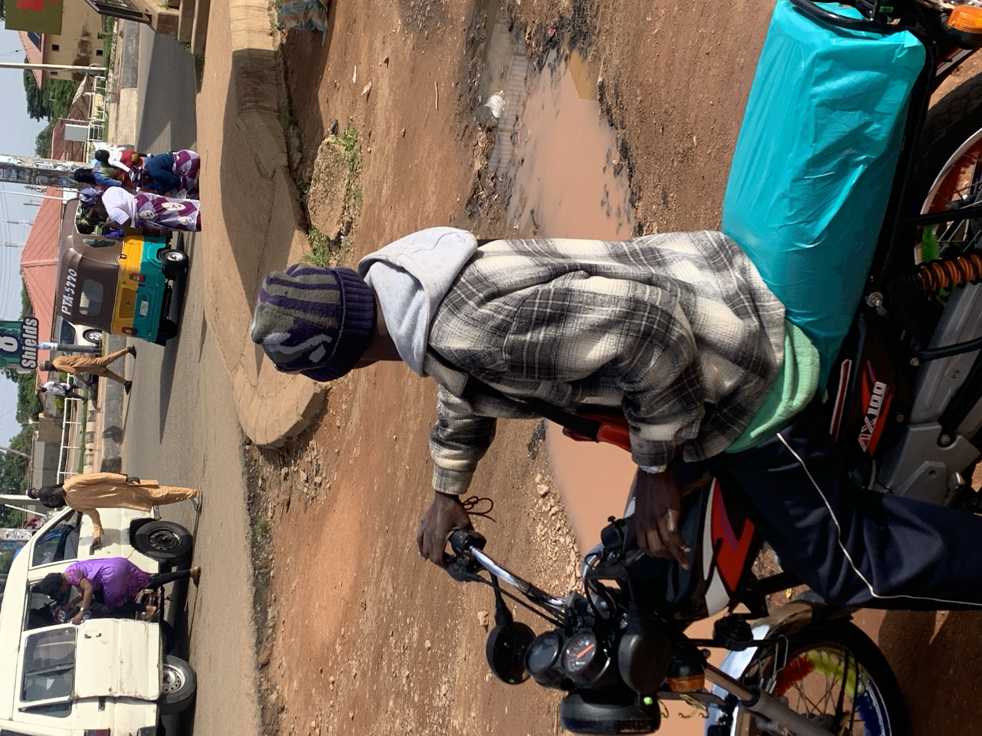 In Gura Top, Nigeria, a man wearing a grey and white checkered jacket, dark blue pants, and a beanie sits on a motorcycle. The motorcycle has a teal-colored cover over the seat. In the background, a white van is parked, and a woman in a purple dress is getting out. A man in an orange outfit stands next to the van. A yellow and teal auto-rickshaw is also visible in the background, with several people gathered around it. The environment consists of dirt roads and some puddles of water.