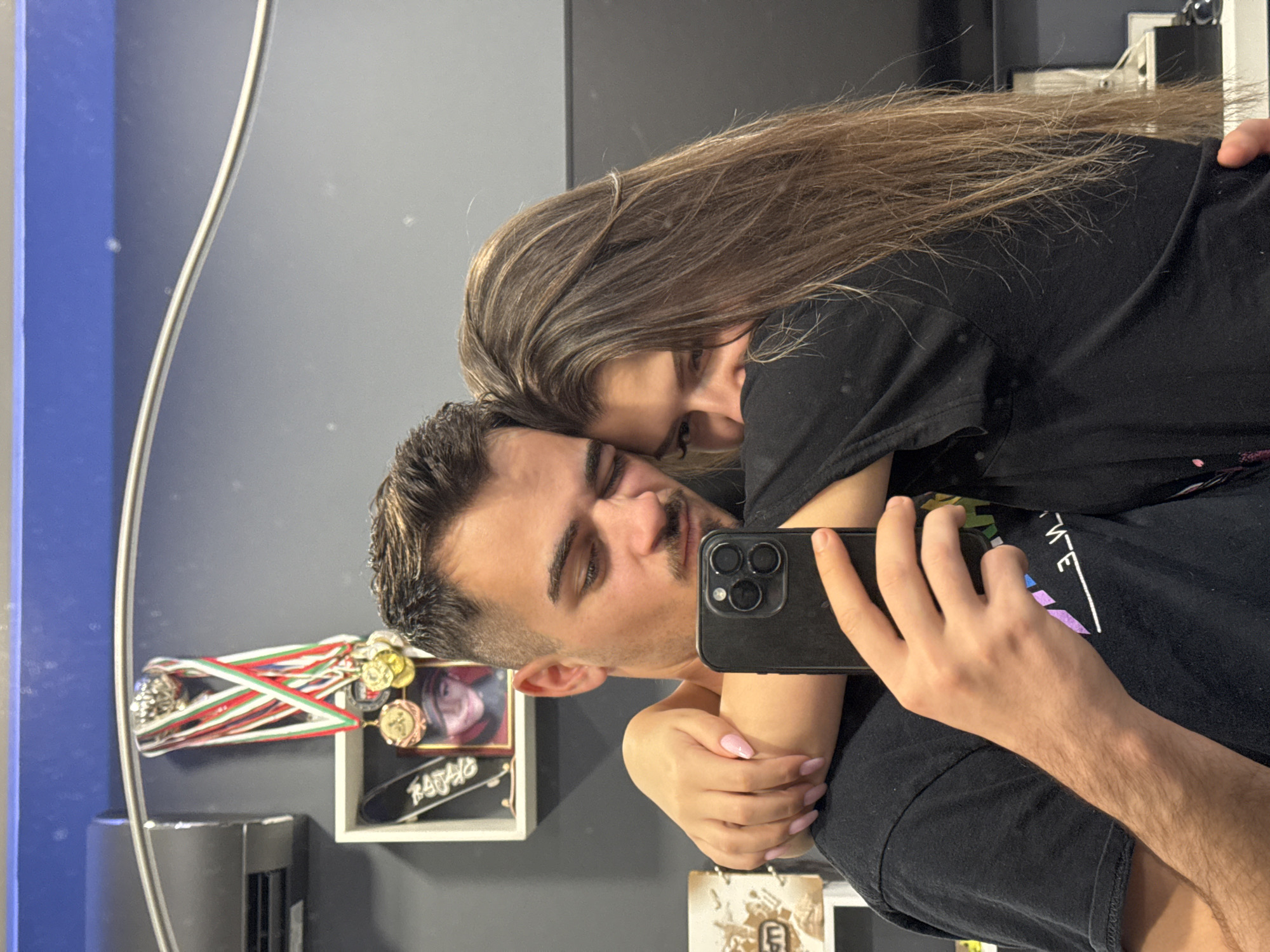 A man and woman are posing for a selfie in a mirror. The man is holding a black smartphone, and the woman is embracing him. Both are wearing black shirts. In the background, a shelf is visible with medals, a photograph, and decorative items. Based on visual clues and the prompt, the location is possibly in Sofia, Bulgaria.