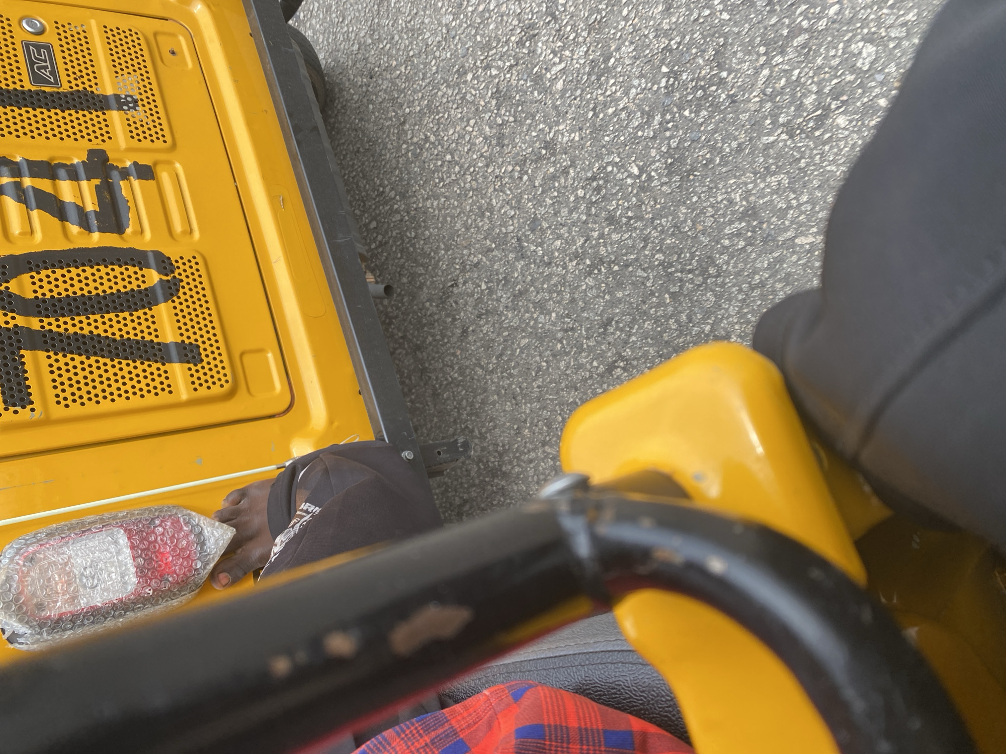 The image shows a close-up view of a person's foot and leg inside a yellow vehicle, possibly a tuk-tuk, on a paved road in Jalingo, Nigeria. The vehicle has the number "7041" painted in black on its yellow body. The person's foot is bare, and they are wearing black pants. A red and blue checkered fabric is visible in the background, likely part of the vehicle's seating. The environment appears to be an urban street, with the rough texture of the asphalt road filling the lower portion of the frame. The vehicle's yellow paint shows some wear and tear.