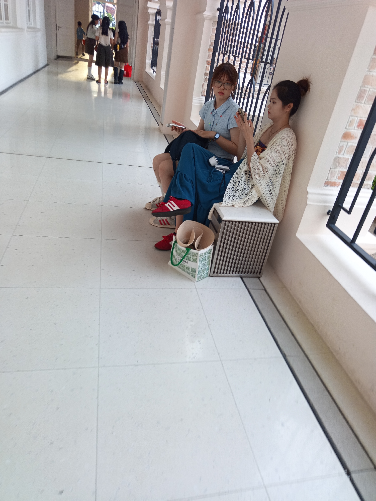 The image captures a bright, tiled corridor, likely within a public building or institution in Kuala Lumpur, Malaysia. The floor is a polished white tile with a subtle speckled pattern, laid out in a grid. Sunlight streams in from the left, casting long shadows and illuminating the pristine floor.
In the mid-ground, three young women stand near a doorway. One, in a dark top and skirt, faces away from the camera. Another, in a white top and grey skirt, looks towards the first woman. The third, in a white top and brown skirt, is partially obscured but appears to be speaking. They are likely students, given their attire.
Further to the right, two other young women are seated. One, wearing a light blue collared shirt and dark pants, has a smartwatch on her left wrist. The other, in a patterned top and a cream-colored, knitted outer garment, holds a mobile phone in her right hand, her thumb poised as if to interact with it. Between them, a small, metallic storage unit with vertical slats is visible. A white bag with green trim and what appears to be rolled-up paper or fabric is placed on the floor near the seated individuals. The wall behind them is a pale cream color, with architectural elements featuring dark, wrought-iron-like detailing and brickwork visible through it, suggesting an older, possibly colonial-era building structure.