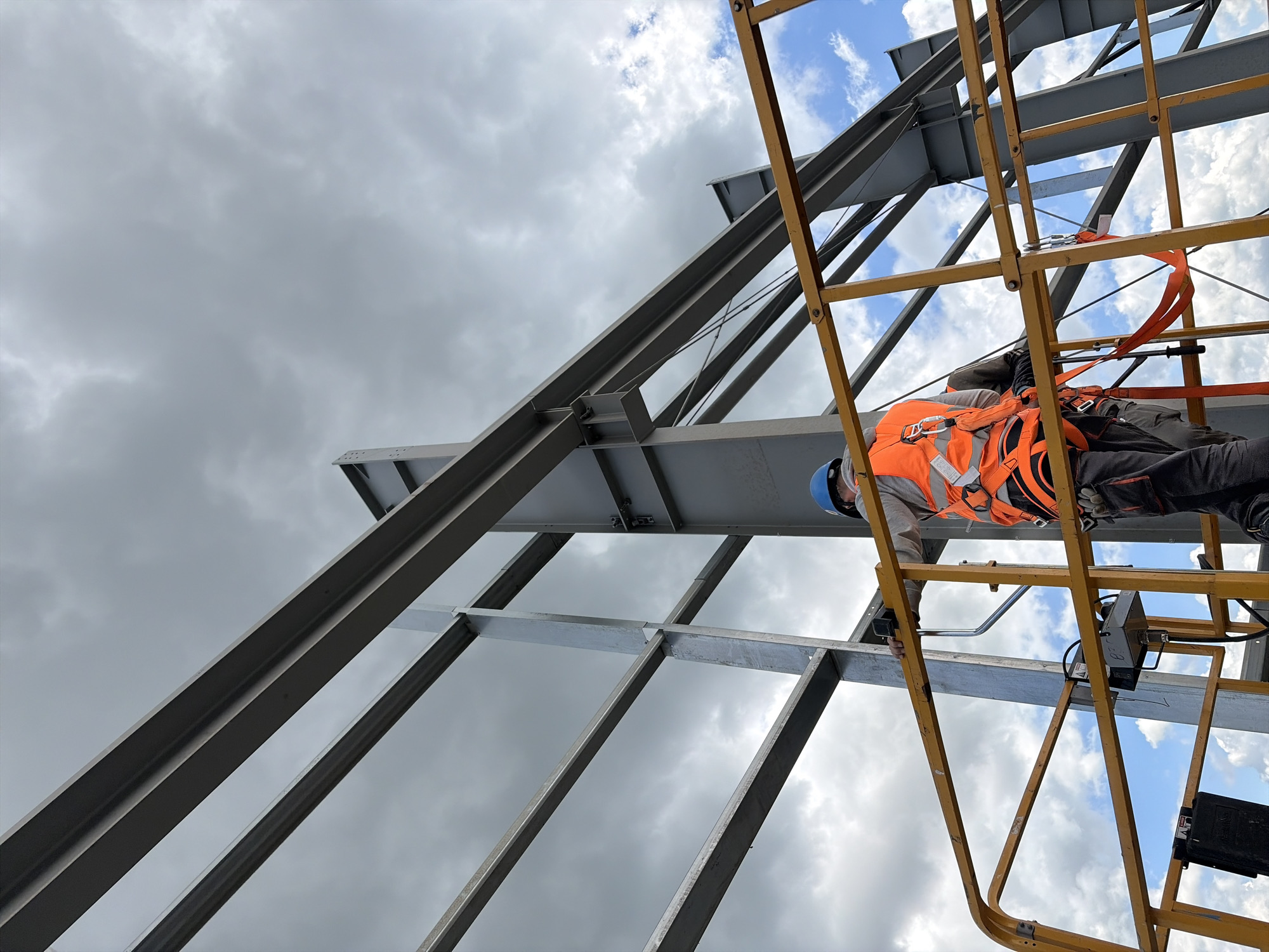 The image shows a construction worker wearing a blue hard hat and an orange safety vest, harnessed and standing on a yellow lift platform. The worker is on a construction site in Stróża, Poland, amidst a metal framework under a cloudy sky. The worker appears to be performing a task related to the construction of a large metal structure, possibly a building or a warehouse. The scene depicts a typical day on a construction site, with elements of heavy industry and personal safety equipment.