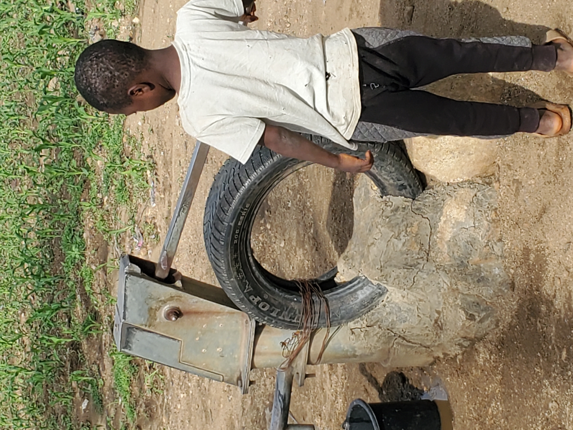 In a rural outdoor setting, a young Black boy stands facing away from the camera, looking towards a water pump. The boy is wearing a tattered white t-shirt and dark pants with gray quilted accents on the sides. He has sandals on his feet. The water pump is made of metal and is attached to a concrete base that has a tire placed around it. The ground is dirt with some scattered stones, and a green crop field is visible in the background. A black bucket, partially filled with water, is situated to the left of the pump. The scene appears to be a functional water source in a dry, perhaps agricultural, environment.