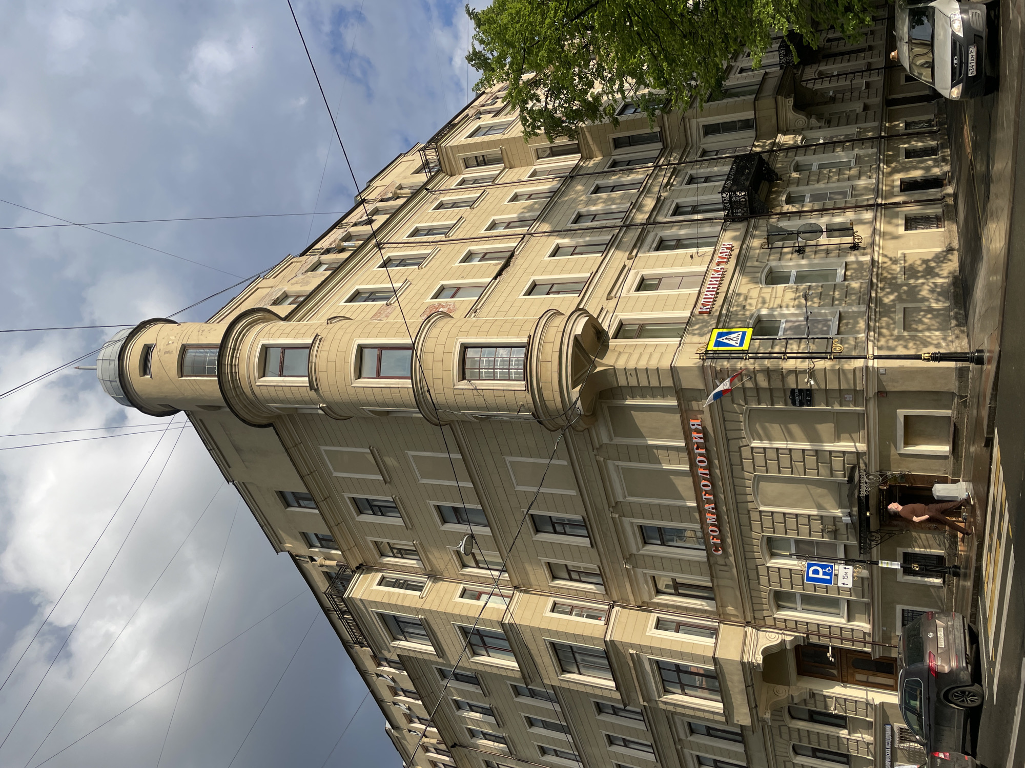An eye-level, wide shot captures a large, multi-story building on a partially cloudy day in Saint Petersburg, Russia. The building, rendered in shades of beige and cream, features a distinctive rounded corner with a series of turret-like bay windows culminating in a glass-domed top. Its facade is adorned with multiple rectangular windows, decorative cornices, and architectural detailing. Green tree foliage is visible near the top right of the building, and utility wires crisscross the sky overhead.
On the street level, a sidewalk runs along the building. A person dressed in a brown outfit stands on the sidewalk, looking towards the building. Several vehicles are parked along the curb, including a light grey Lada Niva Travel (license plate H341AM 157) and a dark sedan.
Prominent red text on the building reads "СТОМАТОЛОГИЯ" (STOMATOLOGIYA), indicating a dental clinic. Higher up, another red sign reads "КЛИНИКА ЛЕН" (KLINIKA LEN), suggesting another clinic. Street signs include a blue parking sign with a "P" and "15M" with an arrow, and a yellow pedestrian crossing sign. Shadows from the sun and trees are cast across the building's facade and the sidewalk, indicating daytime with moderate sunlight.