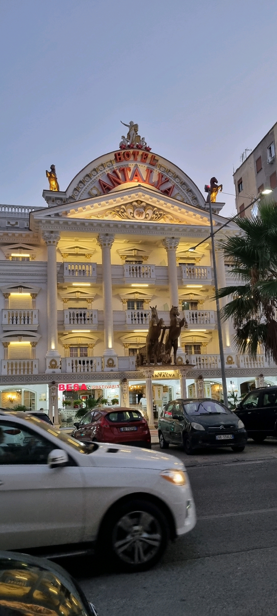 The image depicts the Hotel Antalya in Bashkia Durrës, Albania. The hotel features a grand facade with white columns, ornate gold trim, and a large arched sign displaying "HOTEL ANTALYA" in red lettering. Statues of horses are mounted on the roofline and above the entrance, which is further adorned with a sculptural group of figures. A balcony with a white railing is visible on the second floor. Below the hotel, businesses with illuminated signs are present. Cars are driving on the street in front of the hotel. The sky is a clear twilight blue.