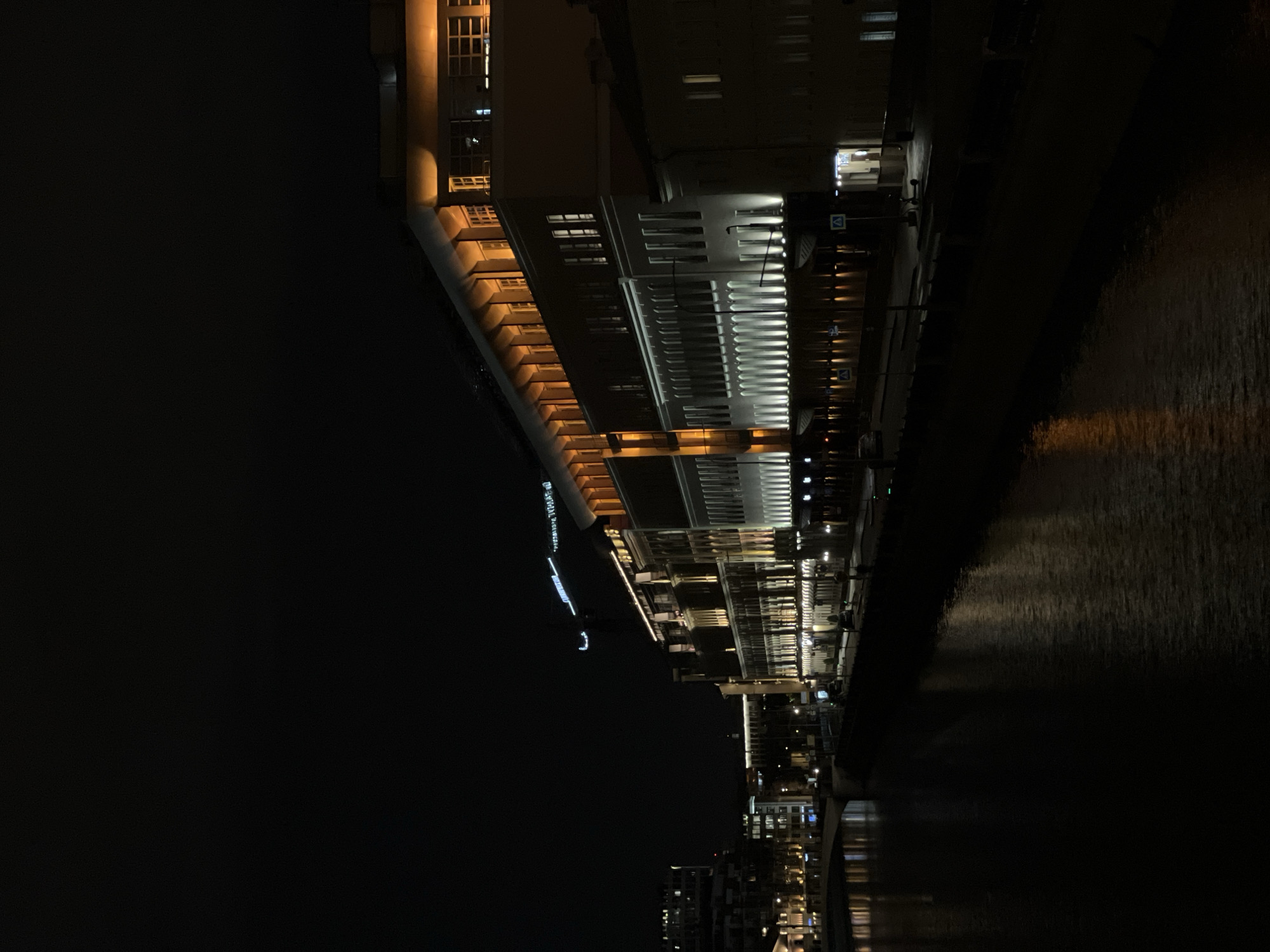 The image shows a nighttime view of the Moskva River in Moscow, Russia. A large building is illuminated along the riverbank, with rows of windows and architectural details visible. The river reflects the lights of the city, creating a shimmering effect on the water's surface. The overall scene is dark with the focus on the lit-up building and reflections in the river. Further in the distance, more buildings and lights can be seen, indicating an urban environment.