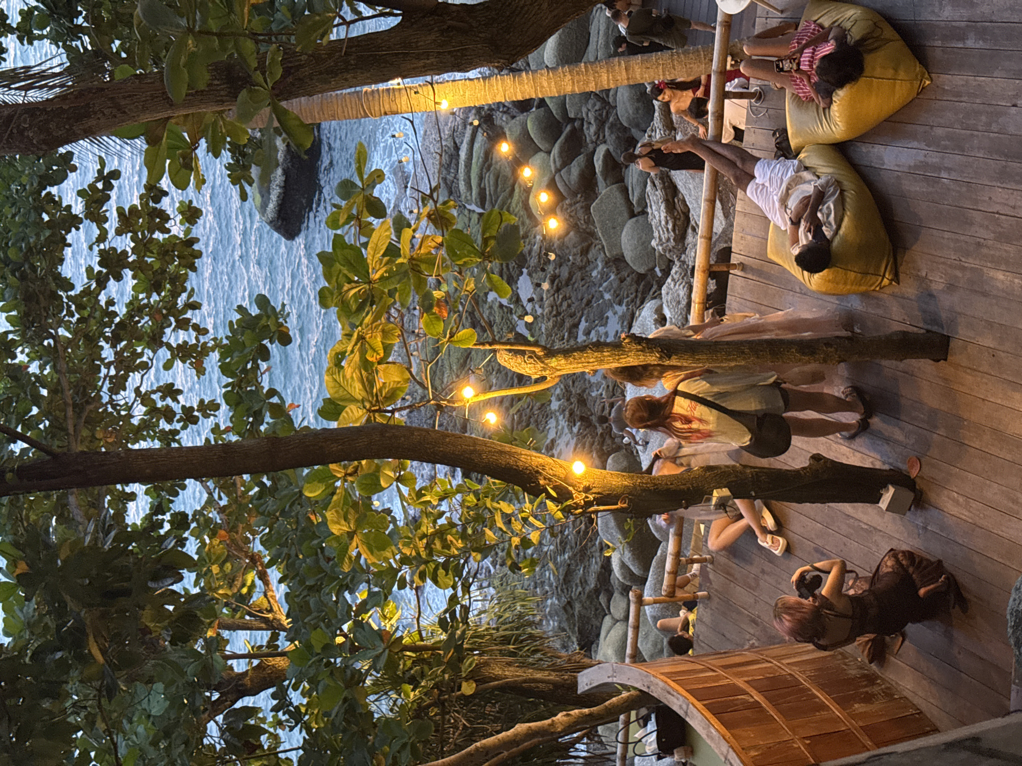 This is an outdoor scene at dusk, likely in Karon, Thailand, featuring a wooden deck overlooking a rocky coastline and the ocean. A group of people are scattered across the deck. In the foreground, a woman with purple-dyed hair kneels and takes a photo with a camera. Further back, several people are seated or lying on large yellow beanbag chairs. One man is lying down with his eyes closed, and a woman next to him is looking at her phone. Other individuals are standing or walking on the deck, some near a wooden railing overlooking the water and rocks. String lights with warm bulbs are hung between trees, illuminating the scene. The ocean is visible in the background, with small waves lapping against the shore and larger rocks present. Lush green foliage from trees overhangs the deck and the view.