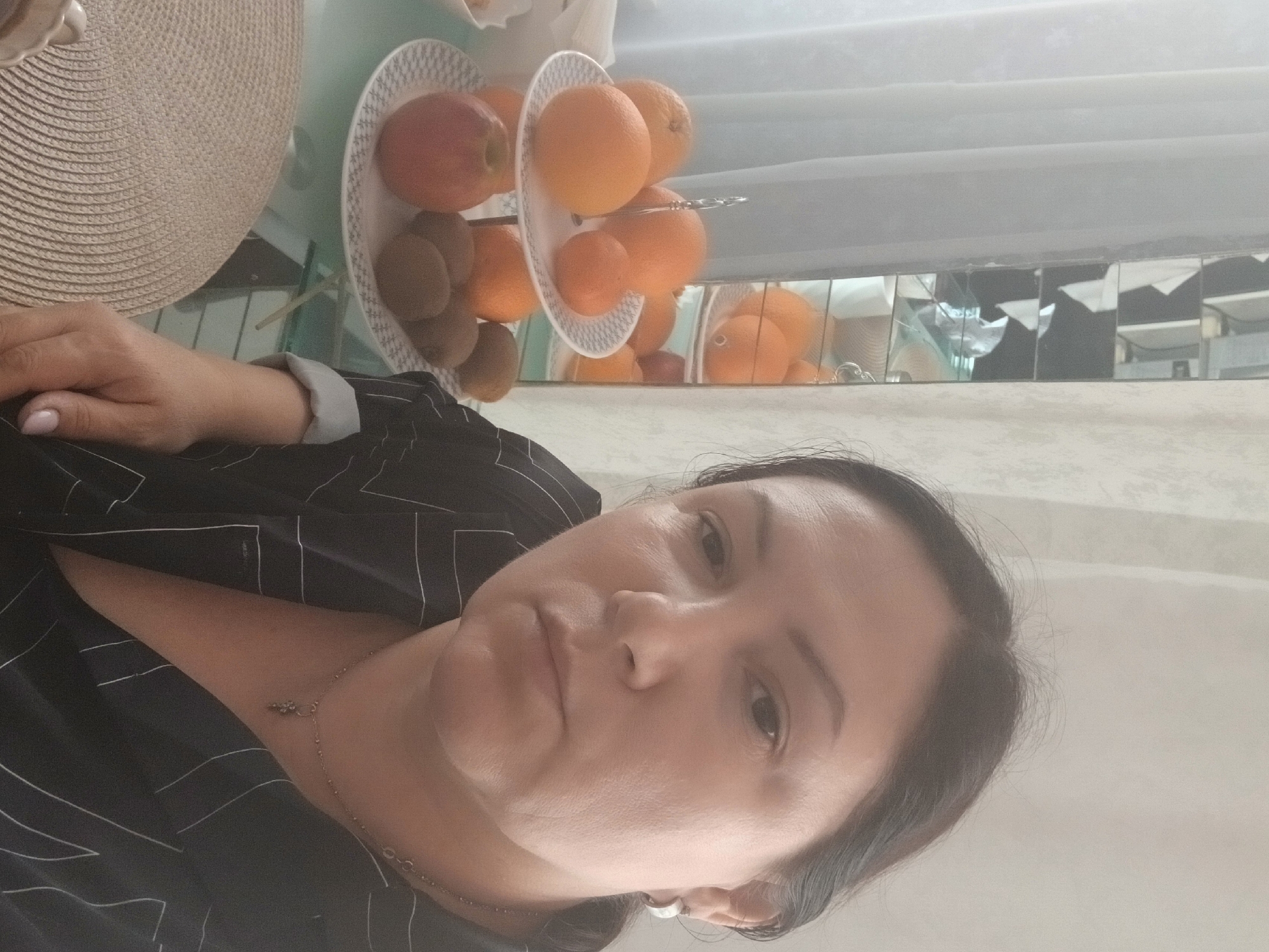 A woman with dark hair and medium skin tone is taking a selfie in what appears to be a dining area, with a table covered by a circular placemat in the foreground. Behind her, a multi-tiered fruit stand holds oranges, kiwis, and an apple. A mirrored cabinet is visible in the background, reflecting a portion of the room. The woman is wearing a black shirt with thin white pinstripes and a necklace. Based on the available metadata, the location is the city of Chaikovsky, Russia.