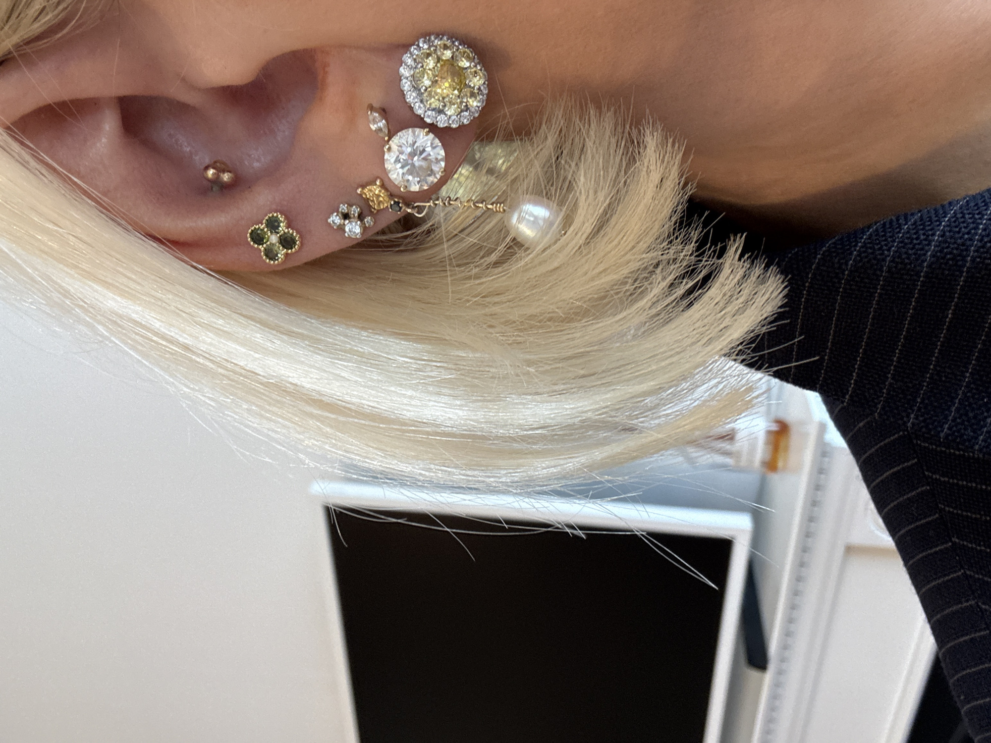 A close-up image captures the right side of an individual's head, primarily focusing on their ear, blonde hair, and the upper shoulder of a dark, pinstriped garment. The ear is extensively adorned with multiple piercings and distinct earrings. A helix piercing contains a small, spherical gold-toned stud. A conch piercing features a gold-toned stud shaped like a four-leaf clover, each leaf set with a dark green stone. Along the earlobe, from superior to inferior, are: a small cluster earring with clear stones; an irregular gold-toned stud; a dangling earring composed of a dark stone, a clear stone, a gold-toned chain, and a baroque white pearl; a large, round, brilliant-cut clear stone stud; and a large, round stud with numerous yellow stones surrounded by a halo of clear stones. The background is out of focus, depicting an indoor environment with a white wall, a dark-screened flat-panel television resting on a white mantelpiece or shelf, and a small amber-colored bottle positioned to the right of the television. The specific location within the city of London, United Kingdom, cannot be determined from the visual information provided.