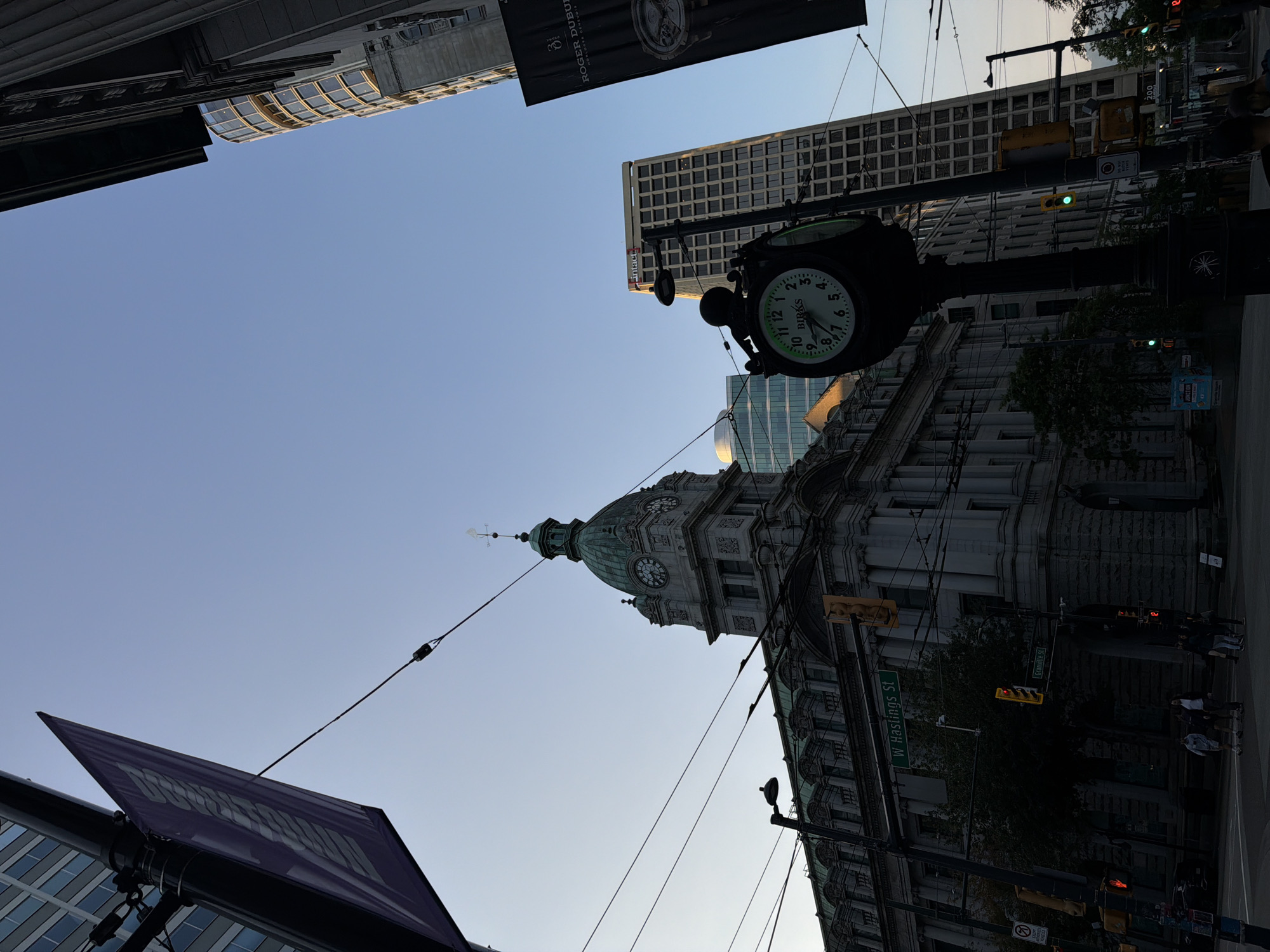 This is a street-level photograph taken in Vancouver, Canada, likely in the Gastown or Downtown area, judging by the architecture and street signs. The image captures an intersection under a clear blue sky. In the foreground, a large, ornate black lamppost with a clock attached stands prominently. The clock face, with the branding "BIRKS," shows the time as approximately 10:18. The lamppost is surrounded by various street elements, including traffic lights, overhead wires for public transportation (likely streetcars or trolleys), and pedestrians crossing the street.
To the left of the lamppost is a grand, historic grey stone building with a green dome-topped tower. This building features intricate architectural details, including arched windows and ornamental carvings. Above this building, a purple banner with white lettering is suspended from a pole. Adjacent to this building and further into the background, a modern, tall office building with a grid of windows is visible, its facade reflecting the sky.
On the right side of the image, another tall building with a predominantly glass facade rises upwards, its shape slightly curved. A large black banner with gold lettering, featuring the "ROGER DUBUIS" brand, is displayed on the side of one of the buildings. Several people are walking on the sidewalks and crossing the street, dressed in casual attire. The overall impression is a bustling urban environment with a blend of historic and contemporary architecture.