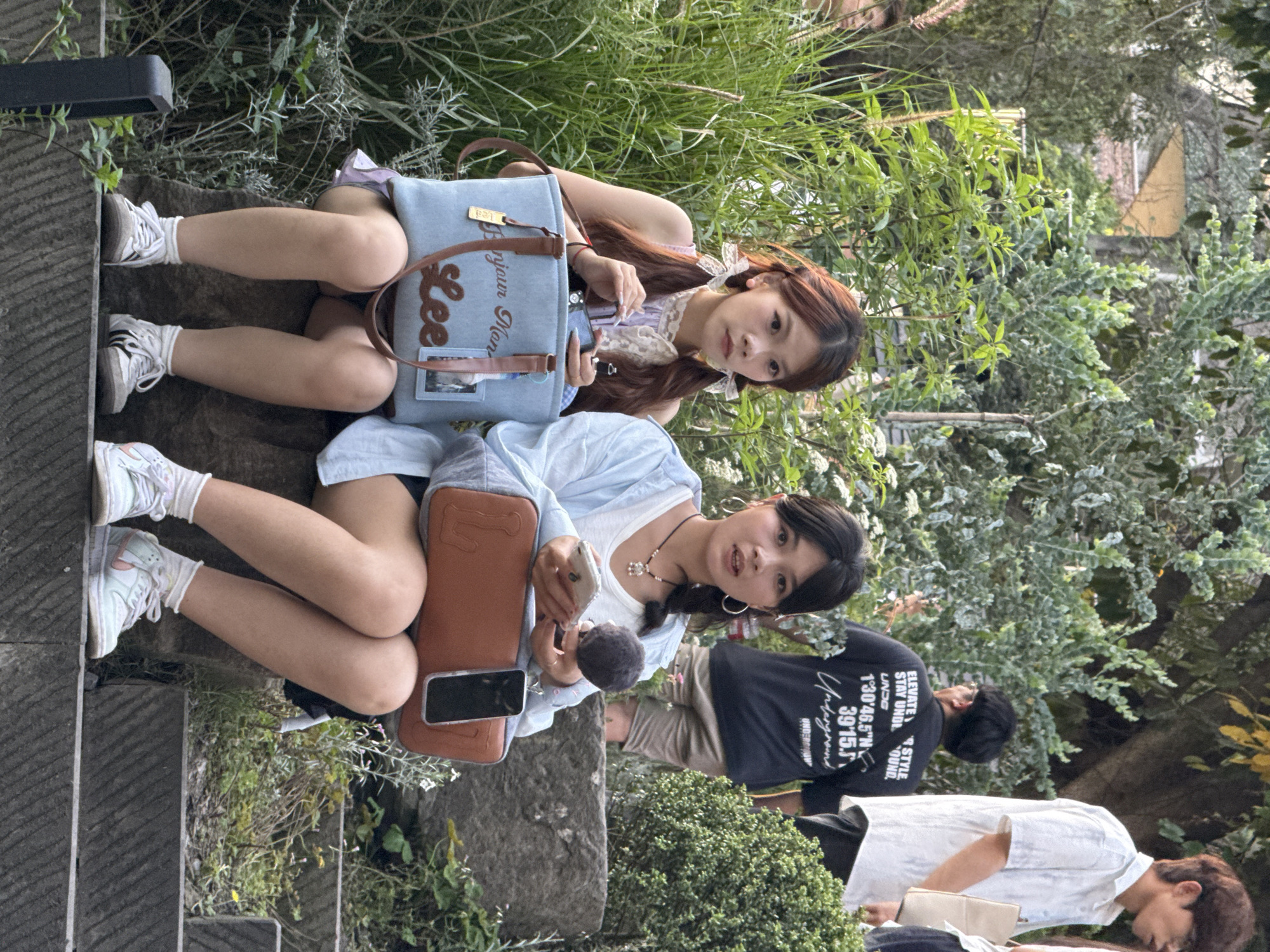Two young women are sitting outdoors. One woman is in the upper part of the image, wearing a light-colored top with a lace collar and pigtails. She has a light blue bag with the name "Lee" on it. The other woman is sitting lower and wearing a light blue jacket over a white top. She is holding a phone and has a brown bag next to her. Both women are wearing sneakers and socks. In the background, there is greenery, including trees and bushes, and other people walking by. Text on a t-shirt worn by one of the people in the background reads "1°30'46.5"N 39'15"E" indicating the location is in Yuzhong District, China.