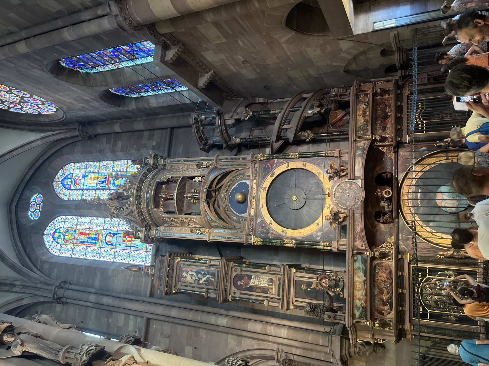 This image captures an interior view of a grand, old building, likely a cathedral or historical church, with a focus on an elaborate astronomical clock. The architecture features high ceilings with stone arches and a series of stained-glass windows, casting colorful light into the space. The clock itself is a massive, multi-tiered structure filled with intricate mechanisms, dials, and decorative figures. A large, dark circular clock face with Roman numerals is prominently displayed, alongside other celestial representations, including a globe and a starry background. Small, detailed paintings and sculptures adorn the clock's various levels, depicting religious or historical scenes.
Several people are visible, mostly clustered around the base of the clock, appearing to admire or photograph it. They are dressed in casual, modern clothing, suggesting a tourist visit. Their expressions seem to convey interest and awe at the complex machinery and the historical significance of the artifact.
The lighting suggests it is daytime, with natural light filtering through the stained-glass windows and possibly some artificial illumination within the building. The overall atmosphere is one of historical grandeur and public fascination. The scene is indoors, within a large, resonant space that hints at significant architectural importance. No specific text is clearly legible enough to be transcribed, though some lettering is visible on the clock's components.