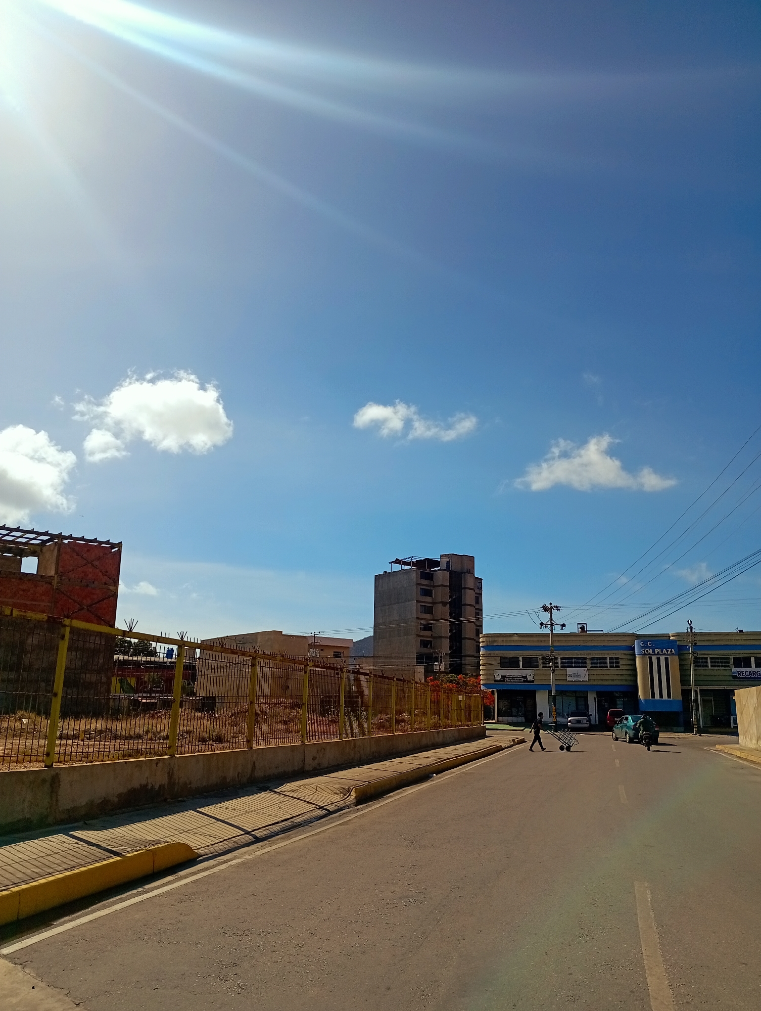 This is a bright, sunlit street scene in Juan Griego, Venezuela. A paved road runs diagonally from the lower left to the upper right of the frame. The sun is high in the bright blue sky, casting strong shadows from a chain-link fence and a yellow curb that runs along the left side of the road. To the left of the road, behind the fence, is a partially constructed building with exposed red brick and a skeletal frame. Further down the street, on the right, is a two-story building with blue and white signage that reads "C.C. SOL PLAZA". Several vehicles are visible on the road, including a green sedan and a motorcycle. A person is walking in the middle of the road, pushing a trolley, and another person is on the back of the motorcycle. In the distance, a multi-story building with a concrete exterior is visible. The overall impression is a typical urban street scene on a clear, sunny day.
