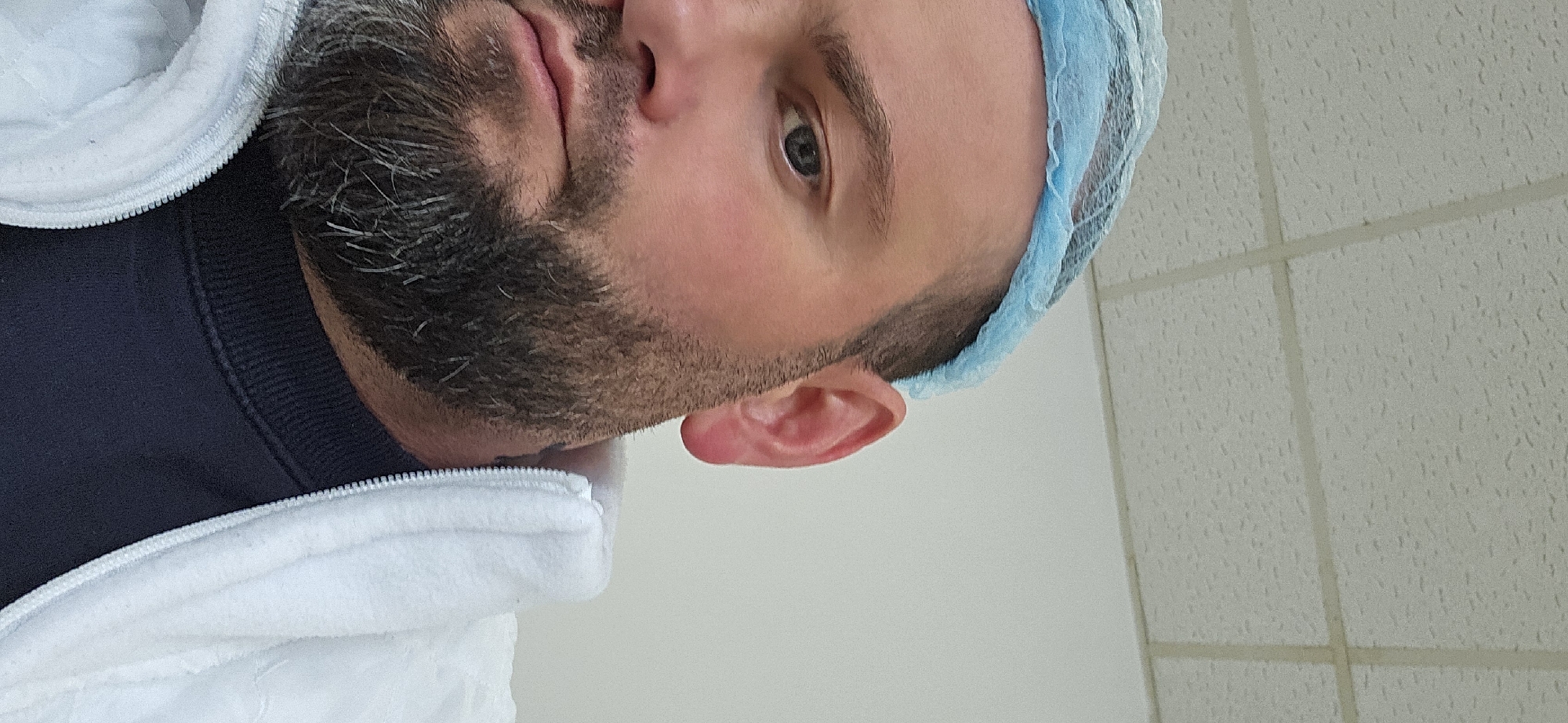 A close-up, slightly angled, portrait of a man with a beard. He is wearing a blue surgical cap and a white coat over a dark blue shirt. The background appears to be a white tiled wall. The location in the city of Delbrück, Germany is not discernable from the image.
