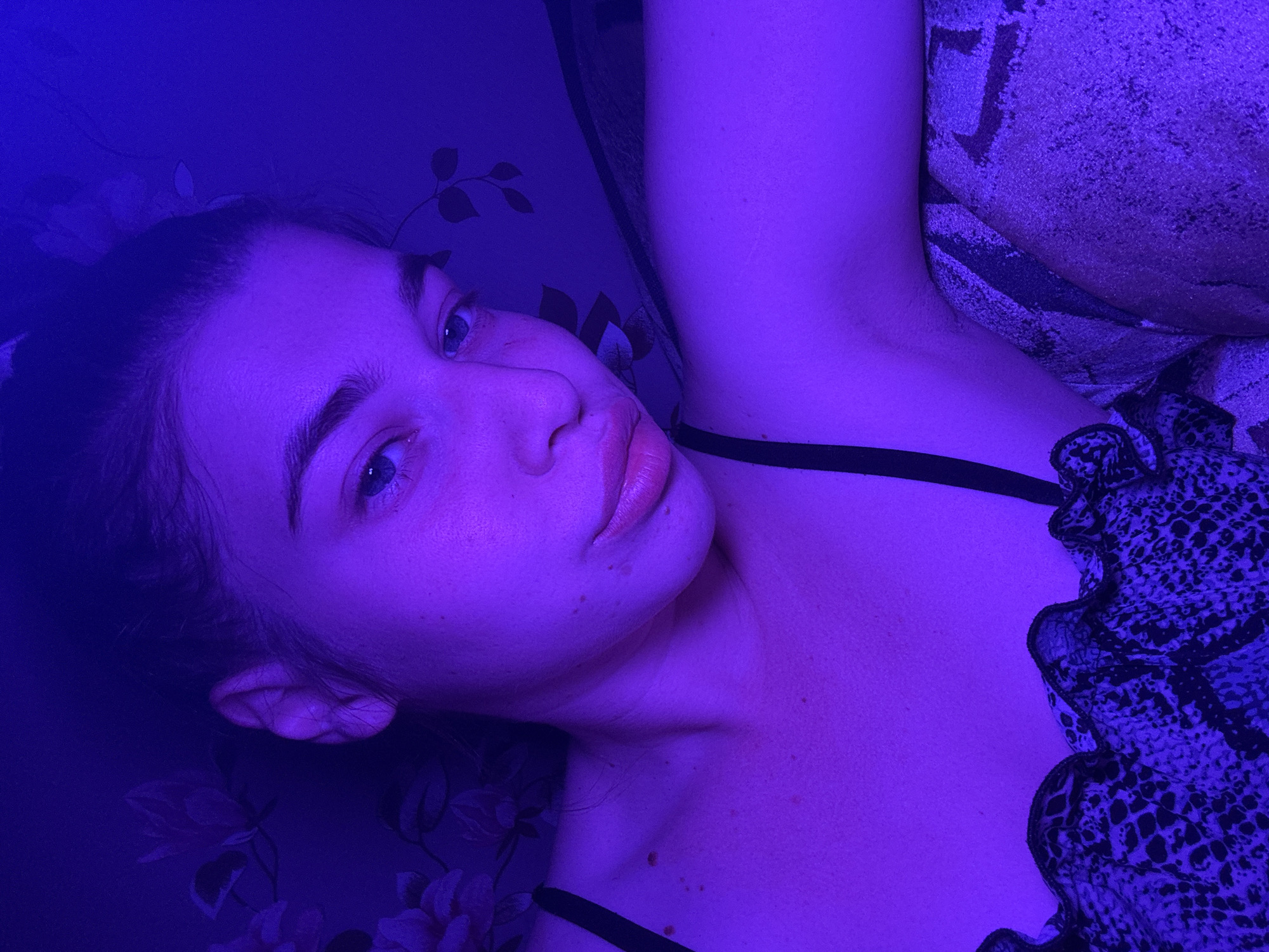 A young woman with light skin and blue eyes is lying down, likely on a bed, with her head tilted towards the left. She has her hair pulled back. She is wearing a top with a black strap and a patterned, ruffled edge. The background is decorated with a floral pattern. The scene is illuminated with a strong blue light. The location in the city of Meshchera, Russia is not discernible from the image.