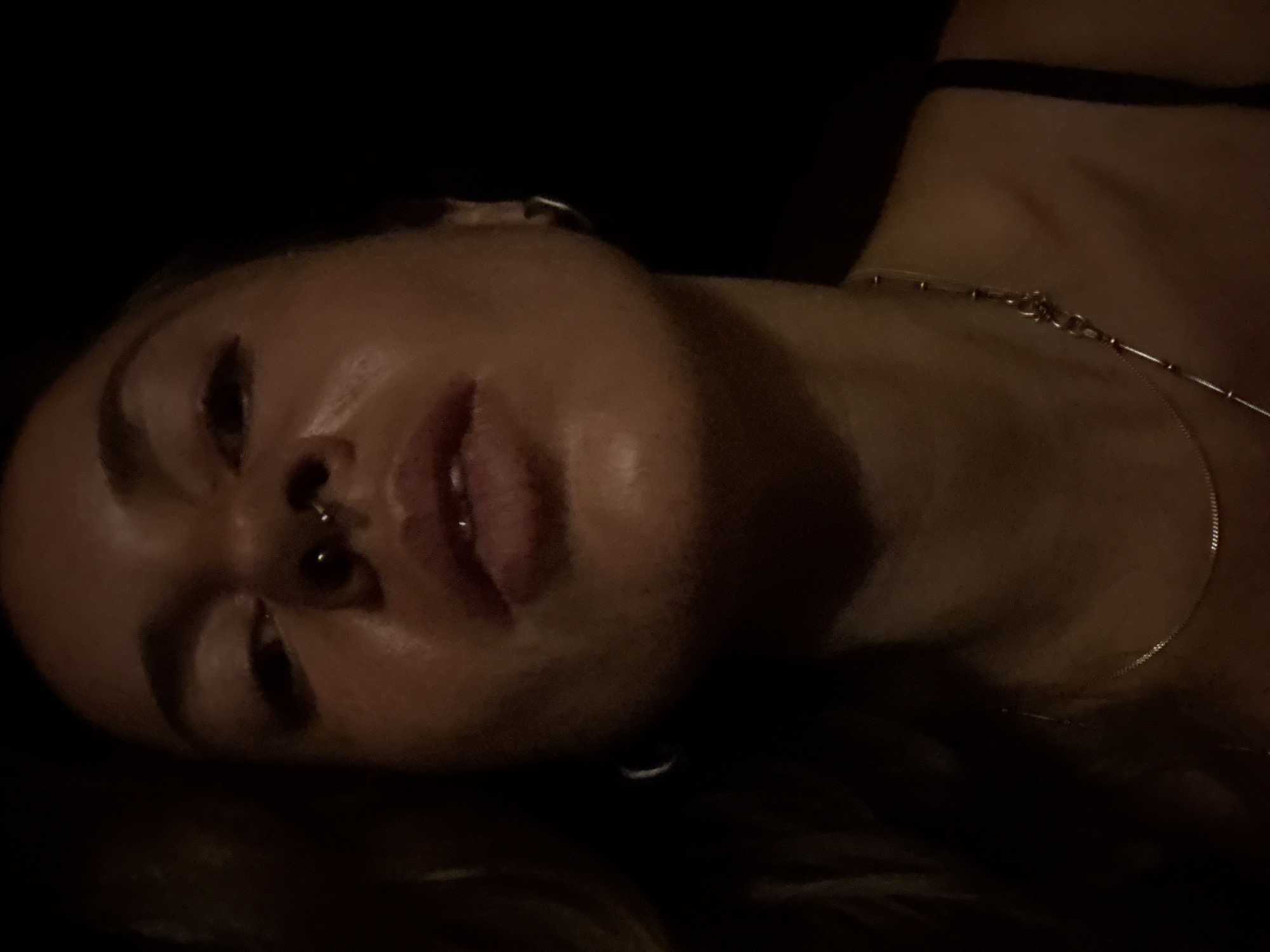 A close-up, low-angle shot shows a woman against a black background. She has a septum piercing and is wearing a necklace. The lighting is dim, and her face is illuminated from above, creating highlights on her forehead and cheeks.