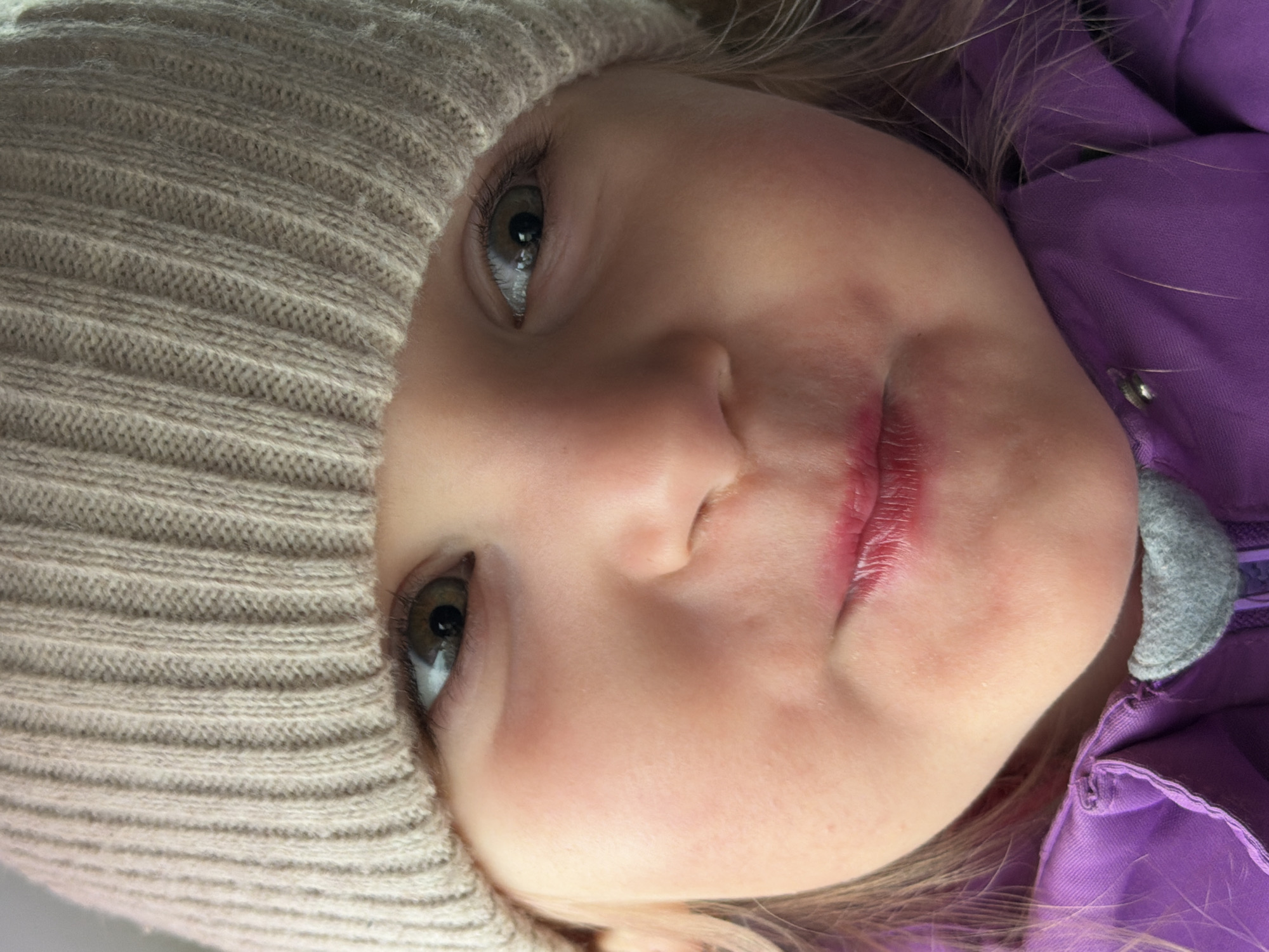 A close-up portrait captures a young child, likely a toddler or pre-schooler, in Chaikovsky, Russia. The image is oriented horizontally, with the child's face filling the frame and tilted roughly 90 degrees clockwise.
The child is wearing a light beige or cream-colored knitted hat that covers a significant portion of their head and cheek. They are also dressed in a vibrant purple jacket or coat, with a visible grey collar or trim, suggesting attire suitable for cooler weather. Strands of light-colored hair, possibly blonde or light brown, are visible near the child's face and collar.
The child's eyes, appearing to be light brown or hazel, are directed upwards and slightly to the viewer's left, conveying a calm, observant, or curious expression. Their lips are slightly parted and show signs of redness or chapping, possibly from cold or dryness. The child's skin appears fair, with a healthy blush on the cheeks.
The background is indistinct and appears to be a plain, light-colored surface, keeping the focus entirely on the child. No specific location cues are visible within the image itself beyond the provided context of Chaikovsky, Russia. There is no visible text. The clothing implies the photo was taken during a colder season, though the exact setting (indoors or outdoors) is not clear.