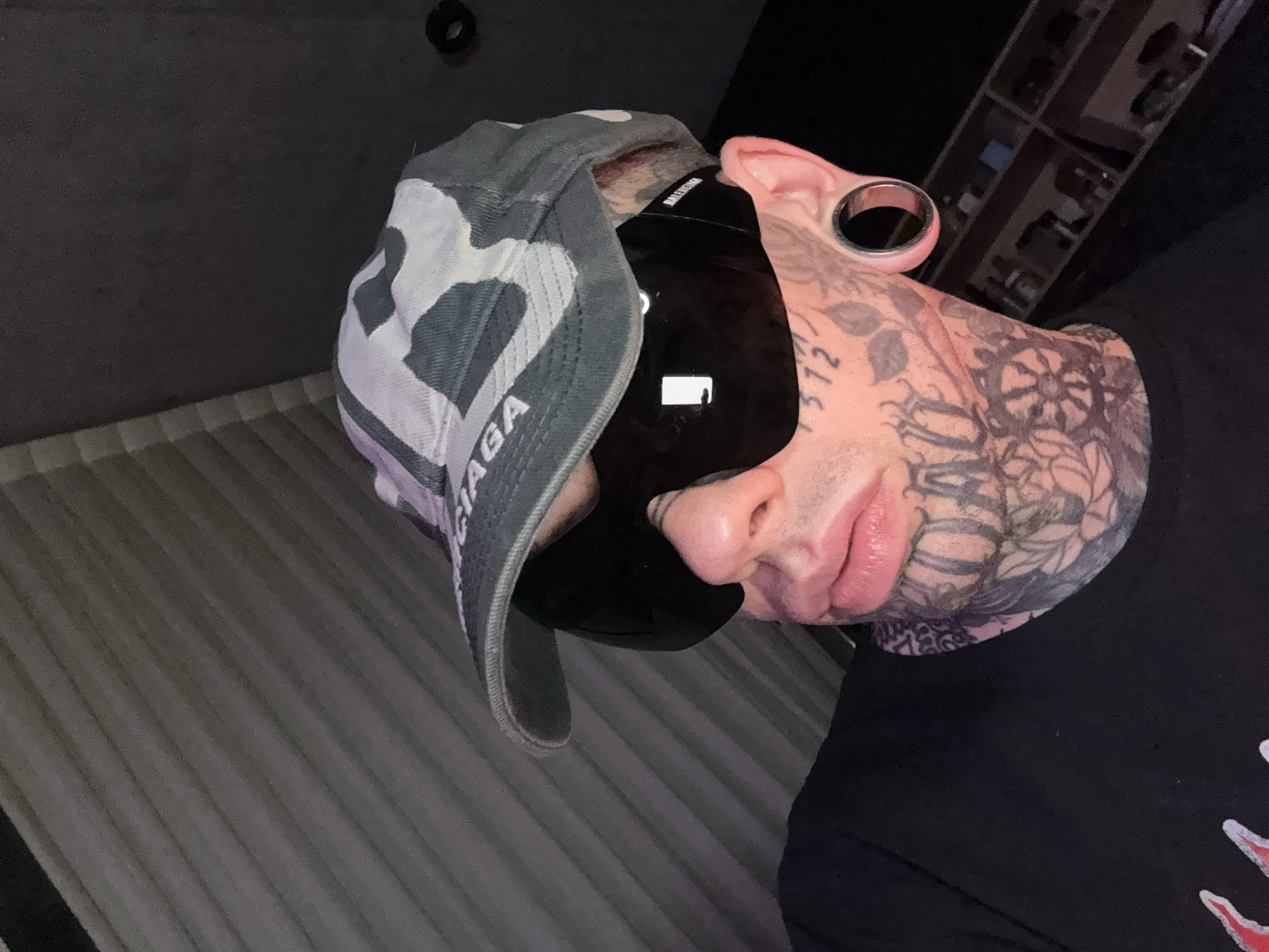 A single individual is depicted in a close-up, dimly lit indoor setting. The subject, appearing male, is shown from the mid-chest upwards, wearing a grey, white, and black camouflage-patterned baseball cap with "BALENCIAGA" branding visible on the front brim. Large, dark, wrap-around Balenciaga sunglasses obscure the eyes, with branding visible on the right temple. The visible skin on the face, neck, and throat is extensively covered with tattoos, including numerical script "372" on the right cheekbone, and various floral and abstract patterns. The lips appear significantly enlarged. The visible right earlobe features a large, metallic, circular tunnel piercing. The individual wears a black t-shirt with a partial red and white graphic design visible on the lower left. The background to the left features a dark, vertically ribbed or textured surface. To the right, a darker, blurred background reveals indistinct shelving units containing various small, dark objects, suggesting a private room or a retail/storage area. Lighting originates from the front-left, casting reflections on the sunglasses. The scene, consistent with an interior space, provides no specific landmarks for a precise location within the city of Buenos Aires, Argentina.