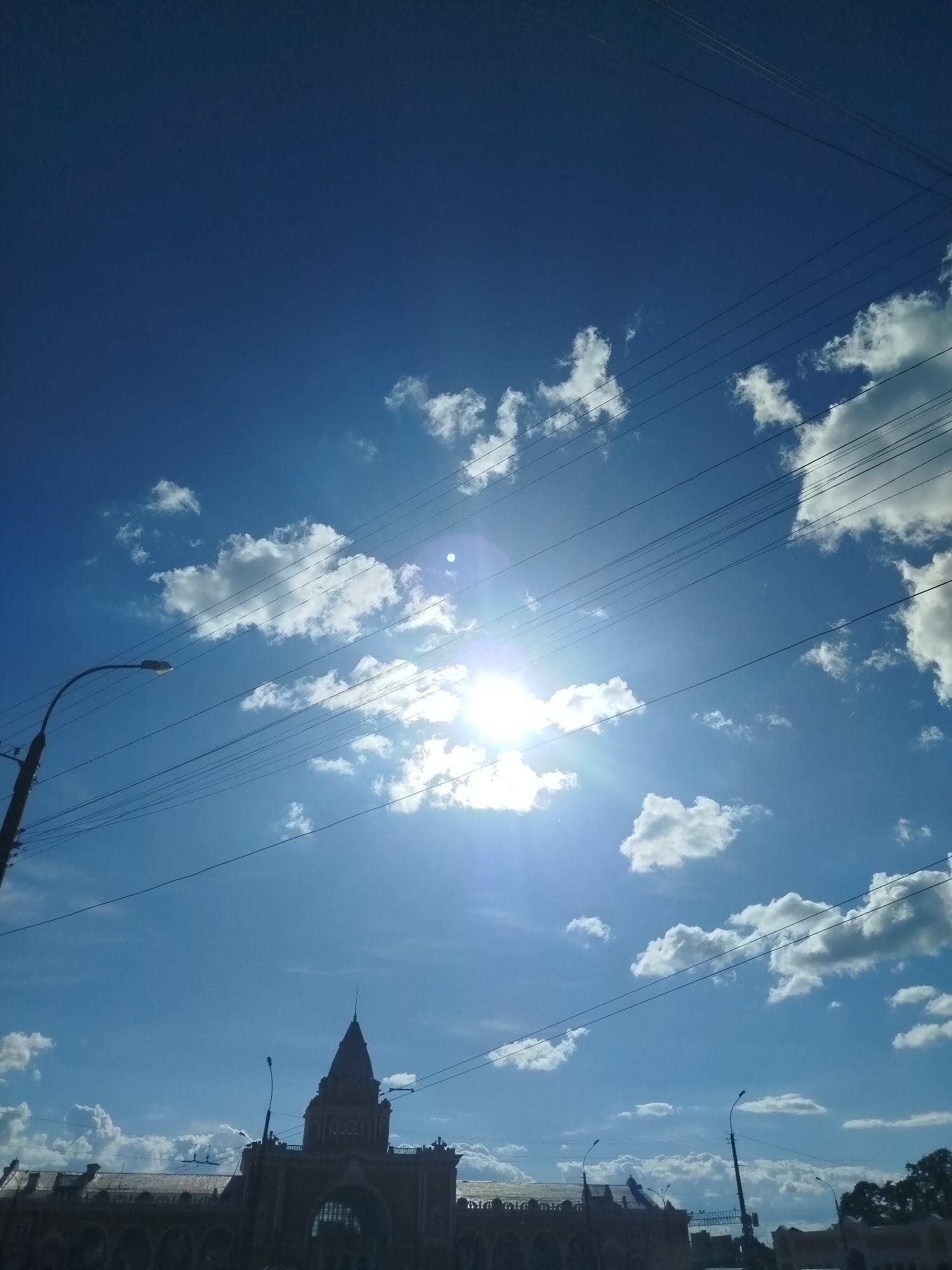 This is a wide-angle, outdoor, daytime photograph taken from a low-angle perspective, looking up towards a bright blue sky with scattered white clouds. The sun is shining intensely, causing lens flare and washing out some of the details in the sky. Overhead, multiple dark utility wires are strung across the frame, creating a crisscrossing pattern against the sky. In the foreground and extending across the bottom of the image is a silhouette of buildings with architectural features that suggest a historical or market hall structure. A prominent central tower with a pointed spire rises above the other rooflines. To the left, a street lamp on a tall pole is visible. The overall impression is that of a sunny day in an urban environment, possibly a city center or a place with significant historical architecture. The image appears to have been taken in Chernihiv, Ukraine.