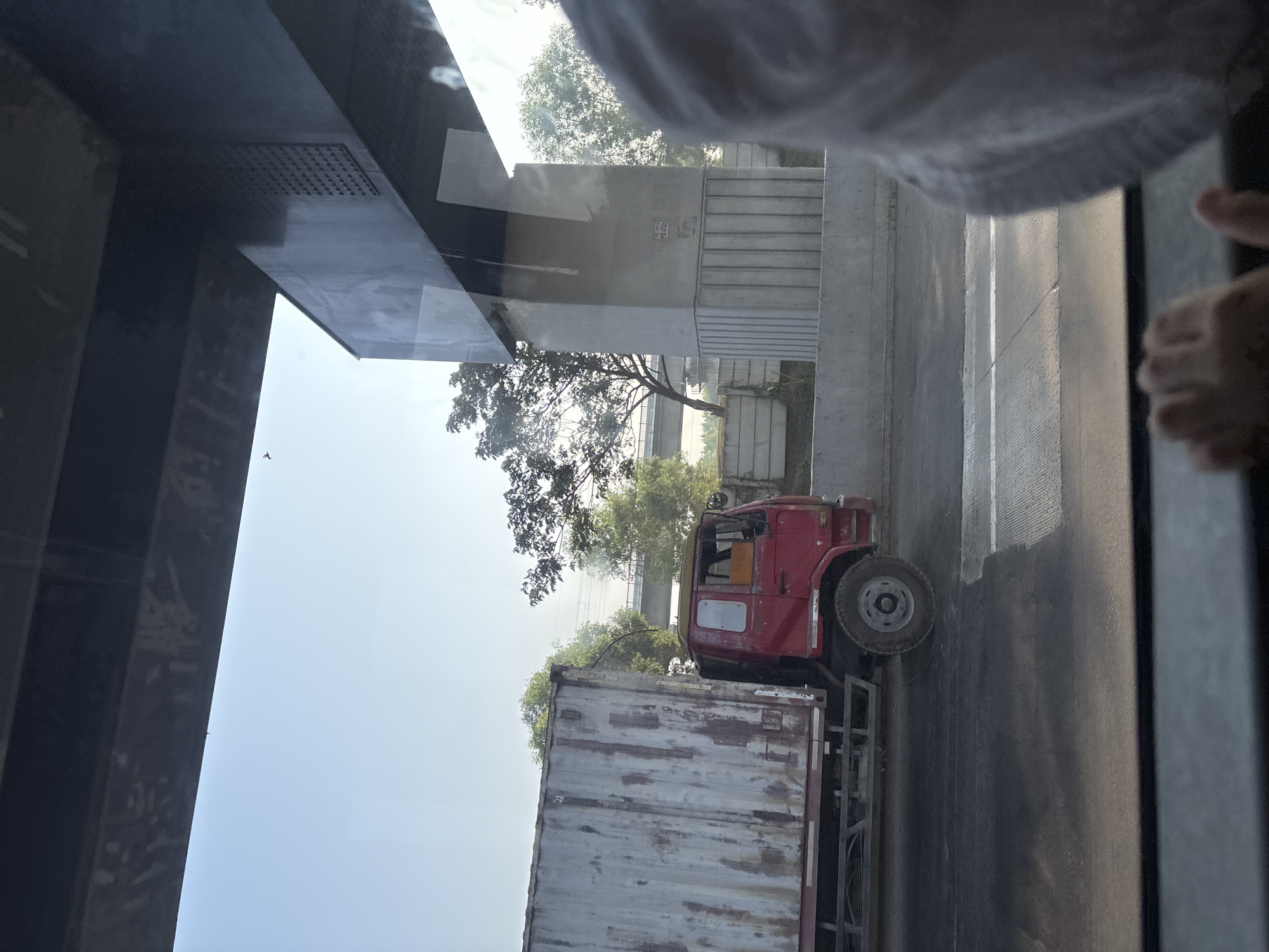 A red truck with a weathered, large silver cargo container is traveling on a highway under a large concrete overpass. The truck is on the right side of the road, moving from left to right. The cargo container is partially obscured by the overpass structure and a reflective surface, likely a window. The scene takes place during the daytime with clear skies. In the foreground, a person's hand is visible resting on a surface, possibly a car window, suggesting the image was taken from inside a moving vehicle. Trees and a bridge are visible in the background, with a concrete wall running along the side of the highway. The overpass appears to be part of an elevated highway system. The location is identified as Hegarmukti, Indonesia.