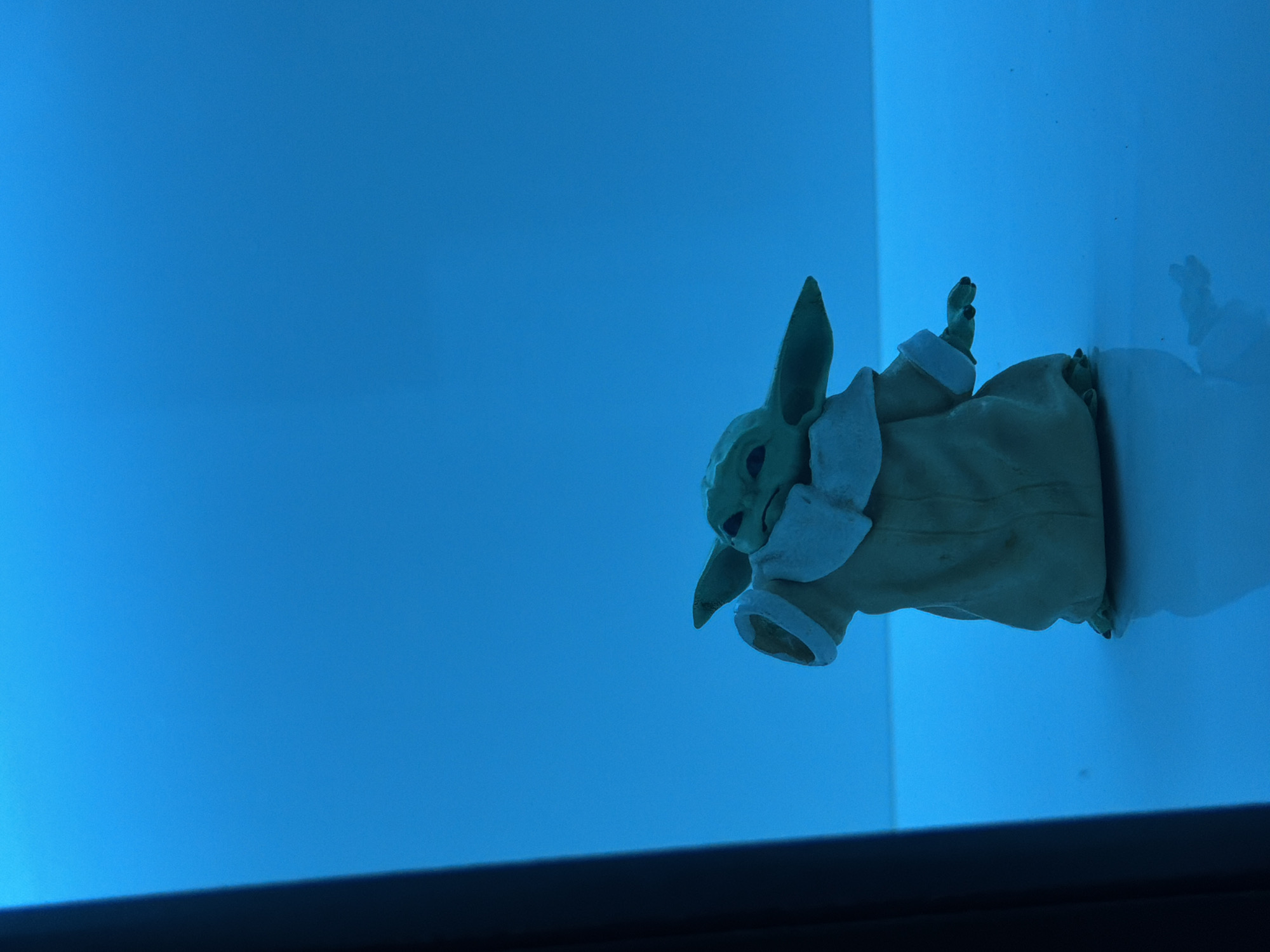 A green, humanoid-shaped figurine, resembling the character Grogu from Star Wars, is positioned in the center of a bright blue, reflective surface. The figurine is wearing a tan robe with a white collar and cuffs. It has large ears and dark eyes. The background is a solid, vibrant blue, with a darker vertical element on the left side of the frame. The lighting is predominantly blue, casting a soft glow on the scene and creating a reflection of the figurine on the surface below. There are no people or specific landmarks visible that would indicate a location in Wonoayu, Indonesia.