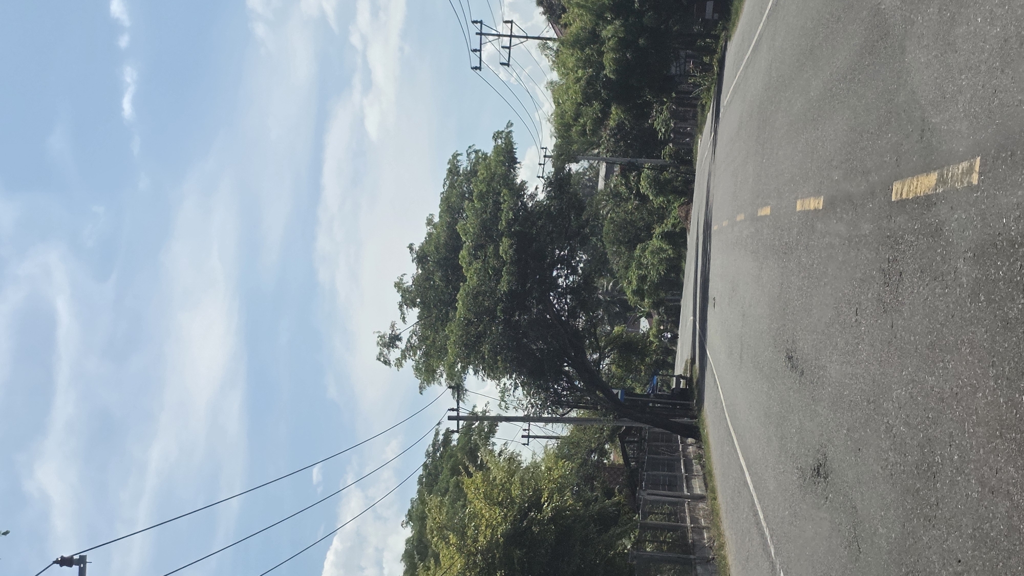 The image depicts a road with yellow dashed lines running along its center, suggesting it is a two-lane road. On the right side of the road, there is vegetation consisting of trees and bushes. Power lines are visible overhead, supported by utility poles. The sky is partly cloudy. The scene appears to be Teungoh Musa, Indonesia.