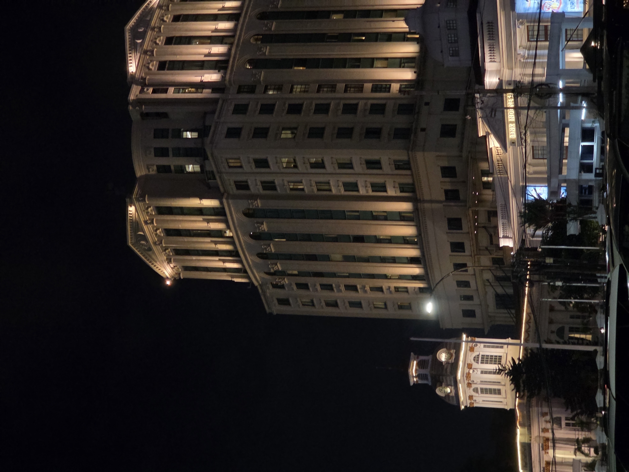 This is a nighttime photograph of a grand, classical-style building, likely a bank, illuminated by artificial lights. The building features multiple floors with numerous windows, pilasters, and decorative moldings, typical of neoclassical architecture. The image is oriented vertically, with the top of the building appearing at the bottom of the frame and vice versa.
The scene is set in a city, with the building's architecture suggesting a historical or prominent public structure. The presence of power lines and streetlights in the background hints at an urban environment. In the lower portion of the frame, a smaller, ornate building with a clock tower is visible, also lit at night. Reflections on a surface, possibly a wet road or a glass structure, are noticeable, adding depth to the scene. The sky is dark, indicating nighttime.
The prominent visible text on the building reads "BANK INDONESIA," clearly identifying it as a branch of the central bank of Indonesia. The overall impression is one of architectural grandeur and civic importance, highlighted by the nighttime illumination. The photograph captures a static scene, with no human activity or dynamic events depicted. The lighting suggests the city is active at night, but the scene itself is peaceful and architectural. The weather appears clear, as there are no visible signs of rain or fog.