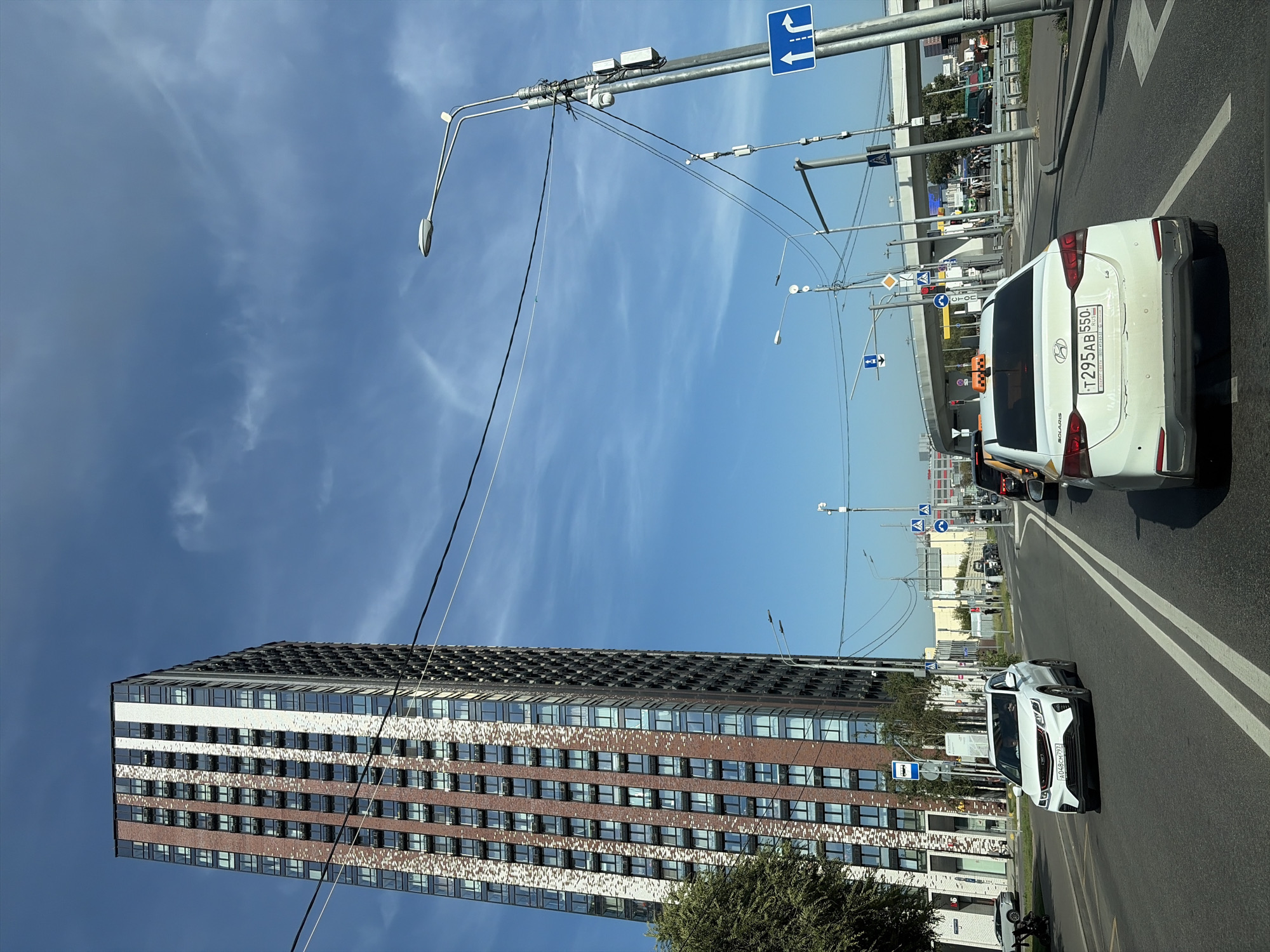 The image captures a street scene in Moscow, Russia, on a clear day with a bright blue sky and scattered clouds. The foreground features a white Hyundai Solaris taxi with the license plate T295AB 550. The taxi has a yellow and orange checkered taxi sign on its roof. To the left of the taxi, a white Geely Coolray SUV is visible, with license plate A048CH 797.
Dominating the left side of the frame is a tall, modern skyscraper with a facade of red brick and white panels, interspersed with numerous windows. The building appears to be a residential complex.
The street is paved with asphalt and marked with double white lines indicating lanes. Several other vehicles are present on the road, including cars, a bus, and what appears to be a truck in the distance. Traffic lights, street lamps, and power lines are visible along the road. Pedestrian crossings are marked on the road with blue and white signs.
In the background, a multi-level overpass or bridge structure can be seen. The general environment suggests an urban setting with significant infrastructure. The presence of a taxi, modern buildings, and traffic indicates an active city environment.