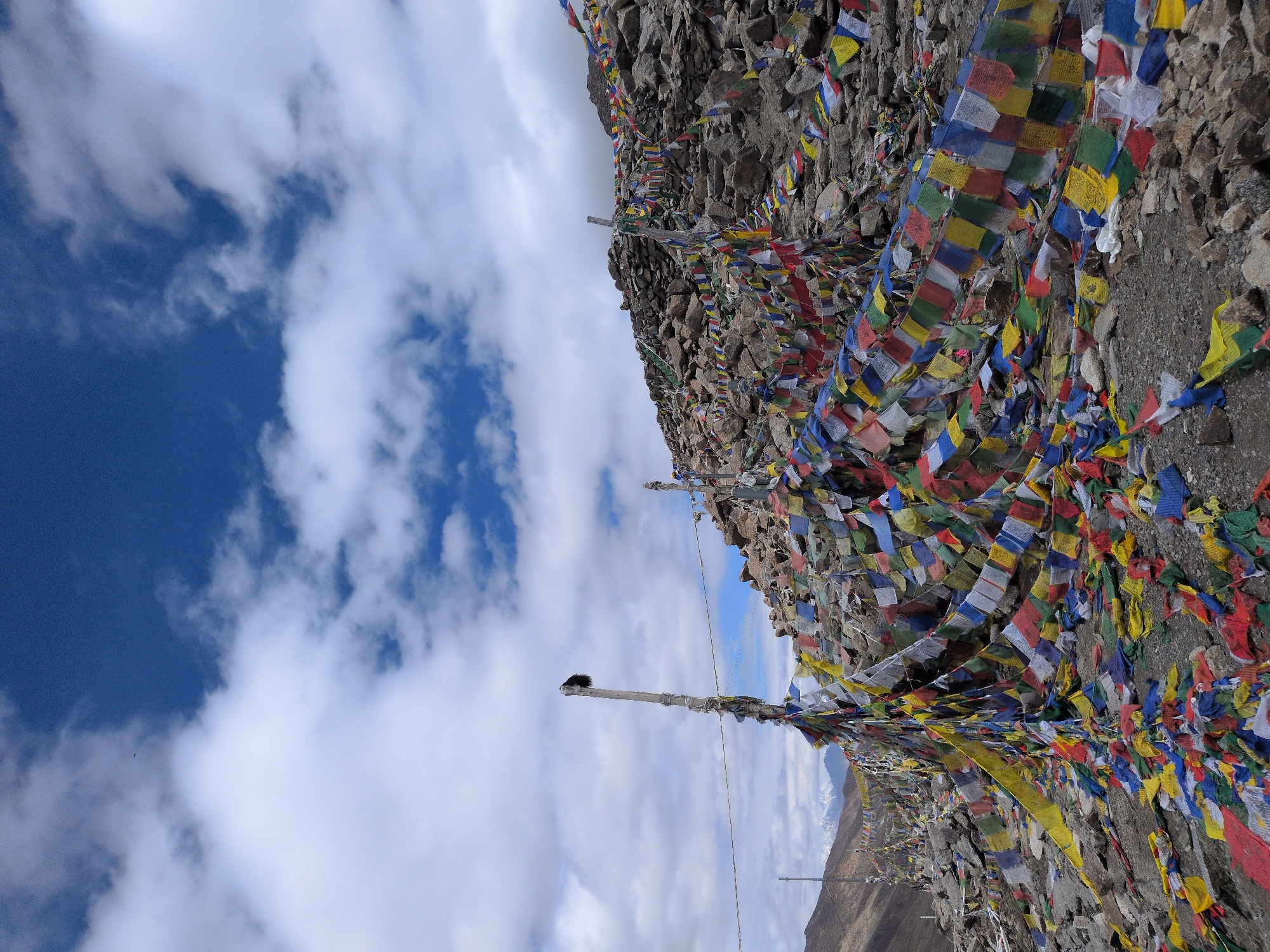 This image depicts a mountainous landscape near Sasoma, India, characterized by a vast expanse of colorful prayer flags. The flags are strung across poles and draped over rocks, creating a vibrant tapestry against the rugged terrain. In the background, snow-capped mountains are visible under a dramatic sky filled with white clouds against a deep blue. The ground is rocky and uneven, with loose stones and gravel. The overall scene suggests a high-altitude, spiritual location.