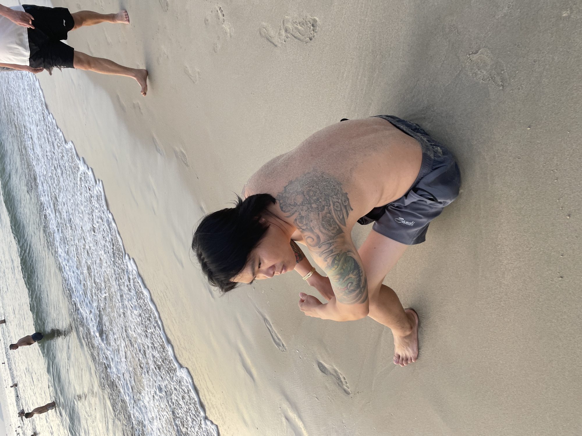 The media file depicts a scene on a sandy beach, likely in Đà Nẵng, Vietnam, given the location context. The image is oriented vertically.
In the foreground, a person with dark hair and tattoos on their arms is crouched down on the sand, looking towards their hands. They are wearing dark swim shorts. To the left, the shallow ocean water is gently washing onto the shore, creating small waves and foam. The beach is sandy with footprints visible in the wet sand near the water's edge and further up on the drier sand.
In the background, to the left and further out in the water, other people are visible, appearing to be standing or wading in the ocean. One person is standing, and another is partially submerged. The lighting suggests it could be late afternoon or early morning, with soft light illuminating the scene. The weather appears to be clear, typical of a beach day.
A notable detail is the text "Sandli" visible on the swim shorts of the person in the foreground. The overall atmosphere appears relaxed, with people enjoying the beach.