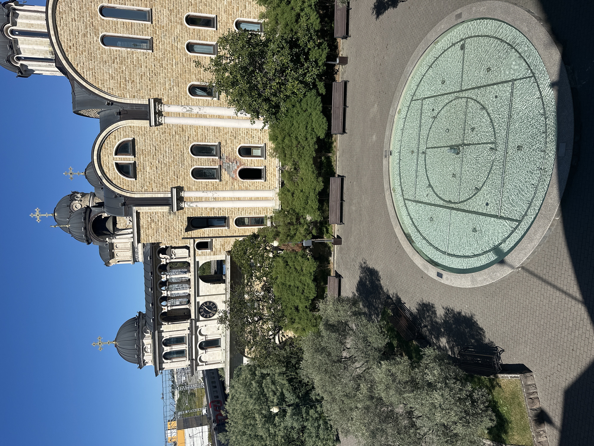 This is an eye-level, high-angle, medium shot of a circular fountain in a city square, with a large building in the background. The fountain is filled with clear, rippling water, and is surrounded by a paved walkway. Several benches are placed around the square, and trees and green spaces are visible. The building in the background is a large, ornate structure made of light-colored stone, with several domes and crosses on its roof, indicating it is a church or religious building. There are arched windows and columns on the facade. A train can be seen passing by in the distance, next to a modern building with yellow accents. The sky is clear and blue, suggesting a sunny day. The location is identified as Sirius, Russia. There are no people visible in the image.
