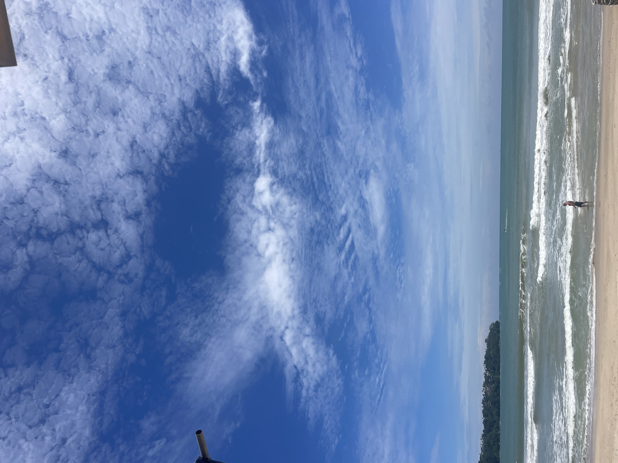 The image depicts a coastal scene in Choeng Thale, Thailand. In the foreground, a sandy beach stretches across the bottom of the frame. A single person, a male, stands partially in the shallow water at the edge of the surf, facing towards the ocean. His attire appears to be dark shorts. The ocean is a deep blue-green, with white-capped waves rolling towards the shore. In the mid-ground, a lush green headland covered in trees juts out into the sea, with some buildings visible amongst the foliage. The horizon line is clear, separating the sea from the sky. The sky is a vibrant blue, dotted with numerous white, wispy clouds that create a textured, patterned effect. A portion of a dark, metallic structure, possibly part of a railing or support, is visible in the upper left corner of the image, intruding into the frame.