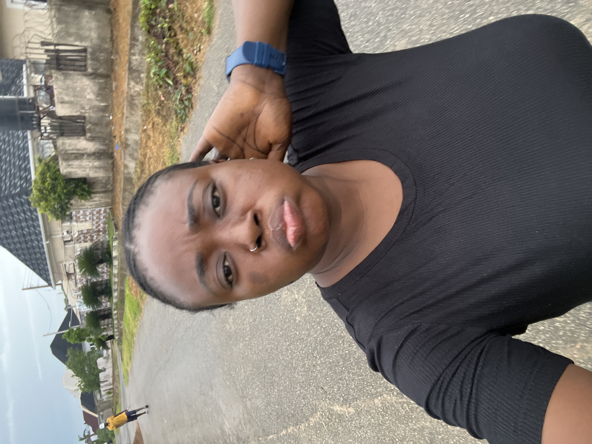 The image captures a selfie of a person, likely a woman, taken outdoors in what appears to be a residential area in Abuja, Nigeria. The photo is rotated 90 degrees clockwise, showing the subject's face and upper body prominently in the foreground.
The subject has dark skin, with her hair styled in braids pulled back from her face. She has a small silver nose ring and is looking directly at the camera with a slightly pouting expression. She is wearing a fitted black ribbed top with short sleeves. A blue digital watch is visible on her left wrist, with her hand positioned near her ear.
The background is out of focus but provides context. It shows a paved street or pathway, possibly asphalt or concrete. Several buildings with various architectural styles are visible, including some with dark roofs and others with textured or unpainted concrete walls, consistent with residential structures in Nigeria. Sparse green vegetation, including bushes and some trees, is scattered throughout the scene. In the distance, a small figure, possibly a child or young person, wearing a yellow shirt and dark shorts, is walking on the street.
The lighting suggests it is daytime, possibly a cloudy or overcast day, as there are no harsh shadows. The overall setting depicts a typical street scene in a Nigerian neighborhood. There is no visible text in the image.