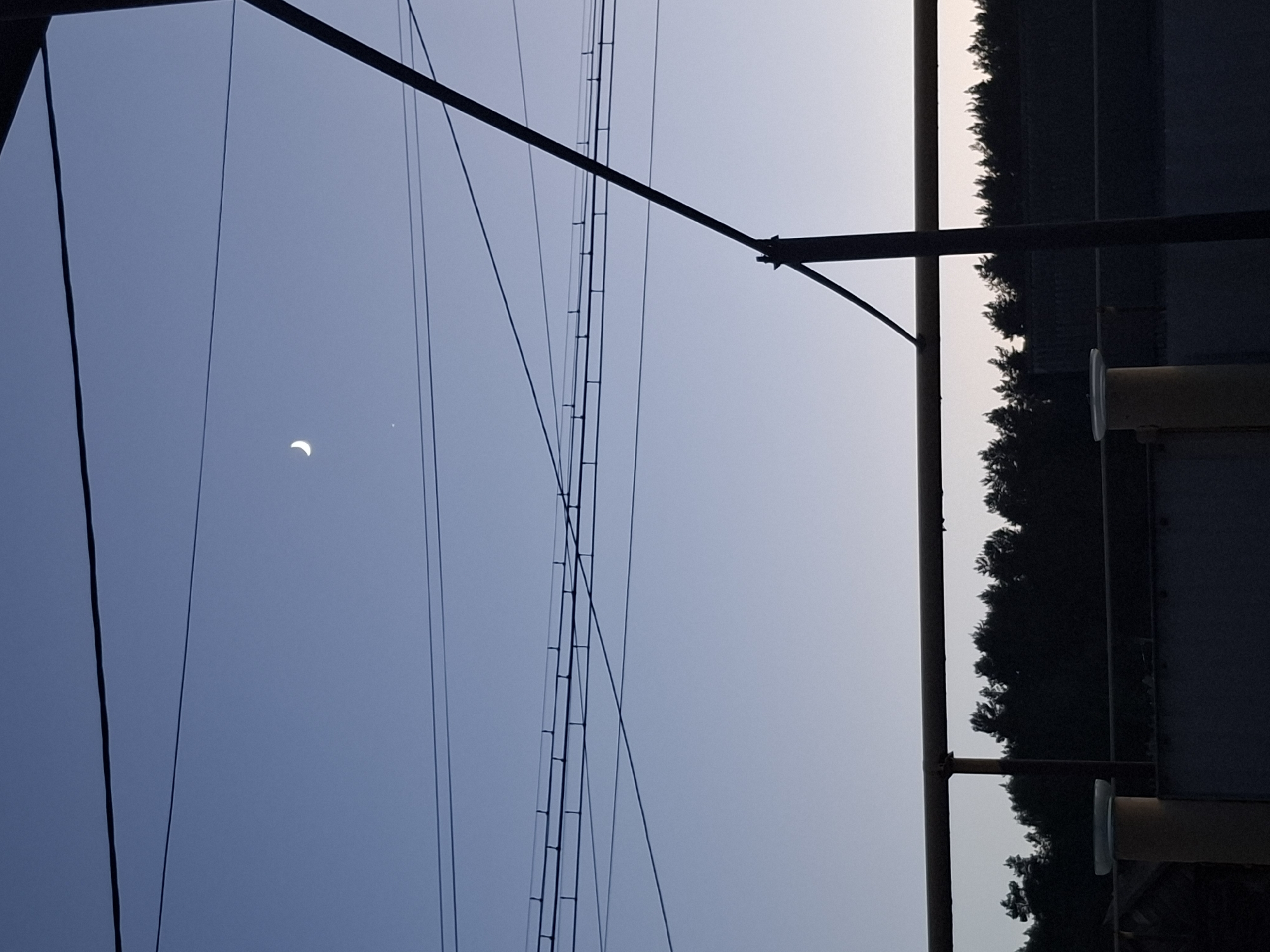 The image, taken in what appears to be Амзя, Russia, shows a partial solar eclipse in a blue sky. Several power lines and a utility pole are visible in the foreground. To the right, a blurred scene suggests a road with trees along the side. The image has been rotated, with the sky on the left and the ground on the right.